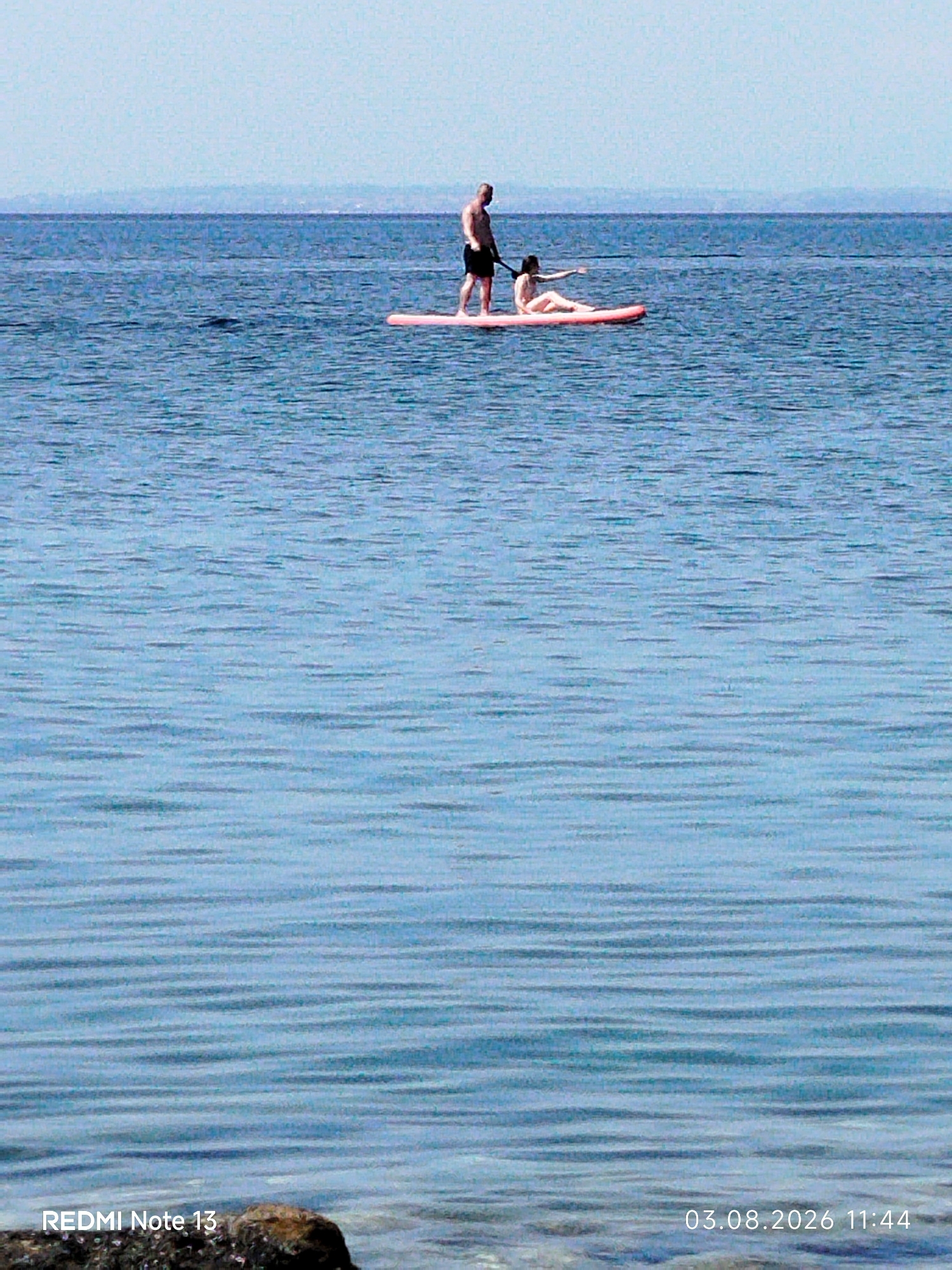 The image shows two people on a paddleboard in the sea. The person in the back is standing and appears to be a male, wearing dark shorts. The person in the front is sitting on the paddleboard and appears to be a female, wearing a light-colored swimsuit. The sea is calm with ripples, and there is a distant shoreline visible. The sky is clear and light blue. The lower portion of the image displays text identifying the device as "REDMI Note 13" and the date and time as "03.08.2026 11:44". The location is stated as Sithonia Municipal Unit, Greece.