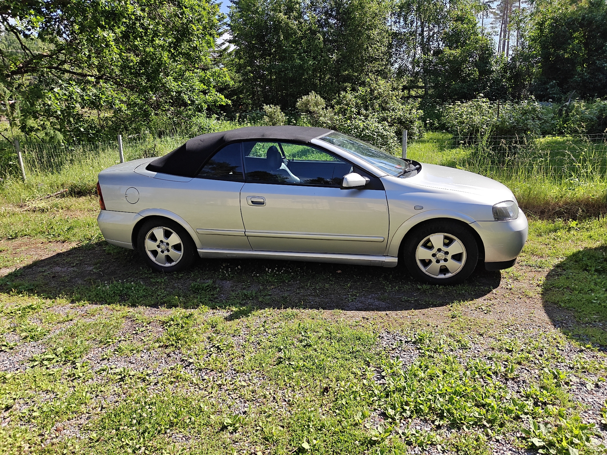 A silver convertible Opel Astra is parked on a gravel and grass area. The car has a black soft top. The scene is outdoors, with trees and a wire fence in the background. The lighting suggests it is daytime and sunny, with some shadows cast on the ground by the car and trees. The car appears to be stationary. The location is identified as Nummela, Finland. There are no people visible in the image. The environment is natural and somewhat overgrown.