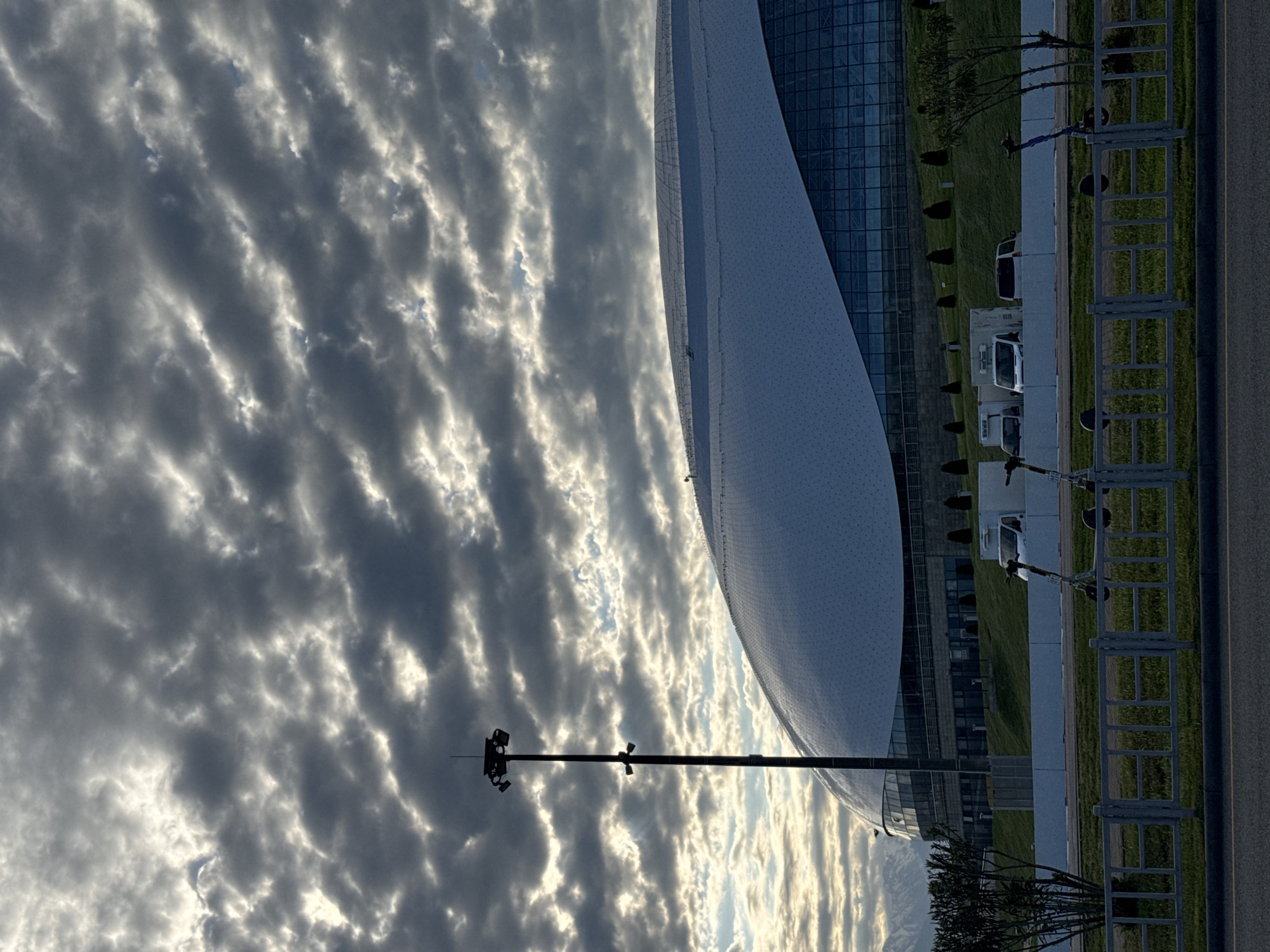 The image, rotated 90 degrees clockwise, presents a modern architectural landscape under a strikingly textured sky, set in Sirius, Russia.
The primary subject is a contemporary building featuring a large, curved facade of white, possibly perforated or tiled material, juxtaposed with extensive dark glass sections that reflect the sky. The building’s design is sleek and futuristic.
Above the building, the sky is filled with dramatic, banded clouds, likely altocumulus or stratocumulus undulatus, appearing in parallel rows. Sunlight breaks through these clouds in places, creating a pattern of bright highlights and deep shadows across the sky.
In the foreground and midground, there is a neatly manicured green lawn, a paved road with a yellow line, and a grey metal barrier or fence structure. Several small, white utility vehicles, possibly electric scooters or similar conveyances, are parked in a row along a white curb. A tall, dark utility pole with multiple floodlights is silhouetted against the bright sky on the left side of the image.
No people are visible. The scene is static, capturing a moment in time rather than an event. The lighting suggests either early morning or late afternoon, with the low sun creating distinct contrasts and shadows, particularly visible in the cloud formations. The weather appears partly cloudy but calm. The modern infrastructure and architecture hint at a well-developed area within Sirius, Russia. No discernible text is clearly visible within the scene itself.
