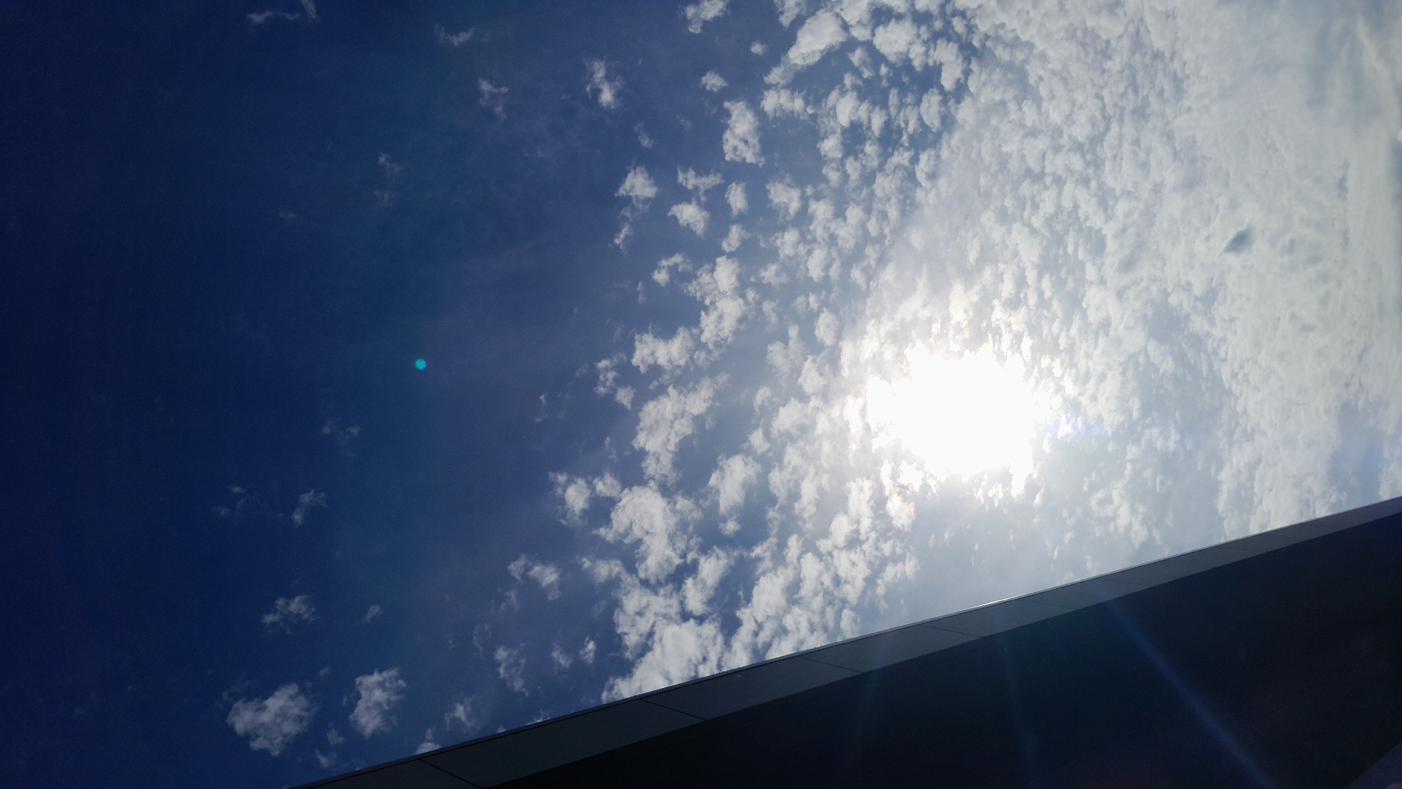 The image shows a view of a bright sun partially obscured by scattered white clouds against a deep blue sky. A dark, angled structure, possibly a building facade or overhang, occupies the lower left portion of the frame, casting a shadow. There are no people or any specific identifiable landmarks to suggest a location in Krakow, Poland. The scene is a natural outdoor environment.