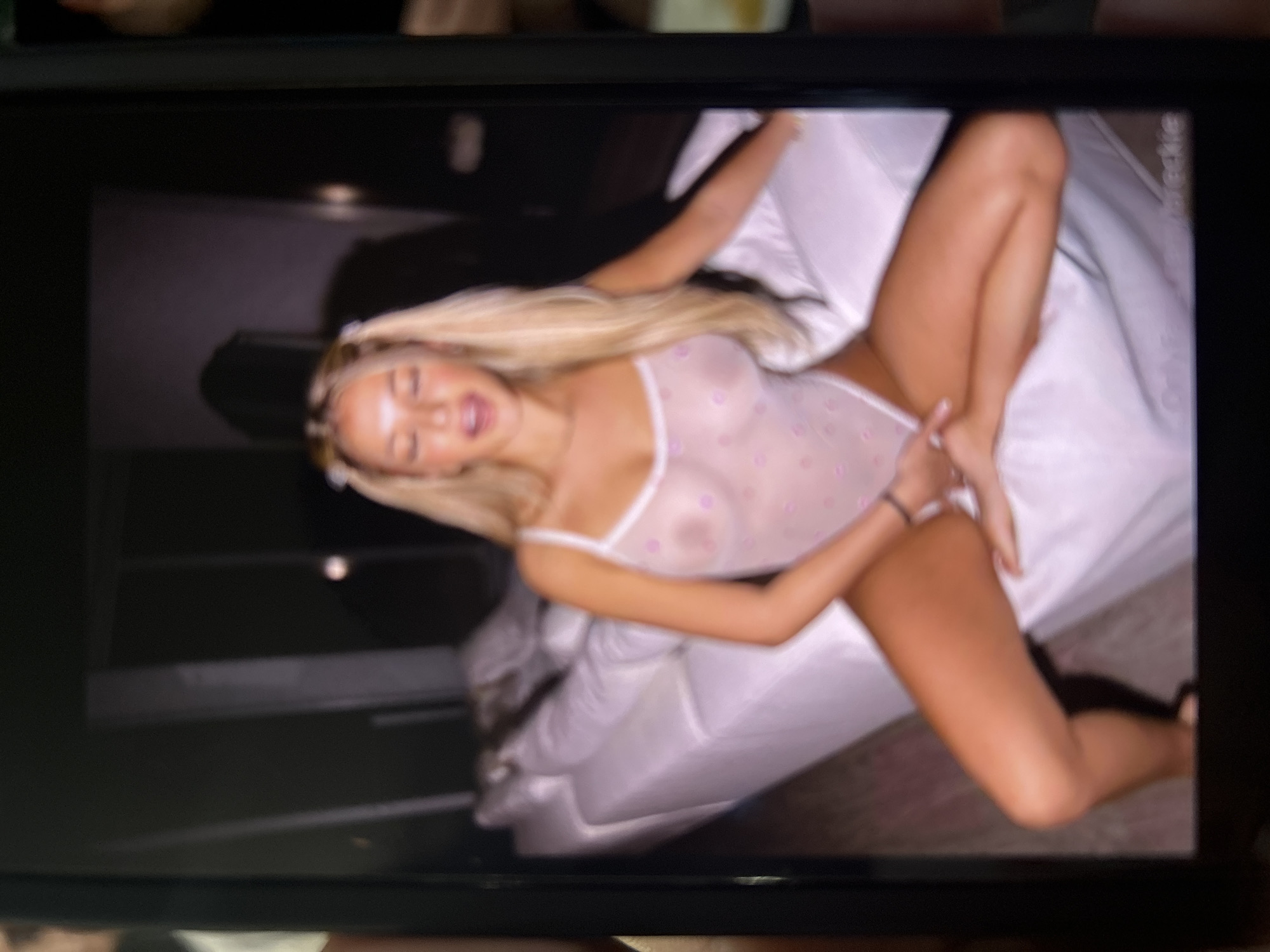 The image displays a photograph of a screen, likely a phone or tablet, exhibiting a horizontally oriented picture. The central subject is a fair-skinned female with long blonde hair styled into two pigtails with light-colored ties. She is wearing a sheer, light-colored (possibly white or pale pink) one-piece bodysuit adorned with small, lighter-colored polka dots, through which her nipples are visible. She is seated on a white sofa or bed, with her legs spread apart and her feet resting on the same surface. Her hands are positioned in her lap, appearing to be touching her private area. Her eyes are partially or fully closed, and her mouth is slightly open. The background is a dimly lit interior space with dark walls and an elongated white sofa or bed extending behind her. Text at the bottom center of the displayed image reads "OnlyFans /breckie". There is no visual information within the image to indicate its location as the city of Blagoevgrad, Bulgaria.