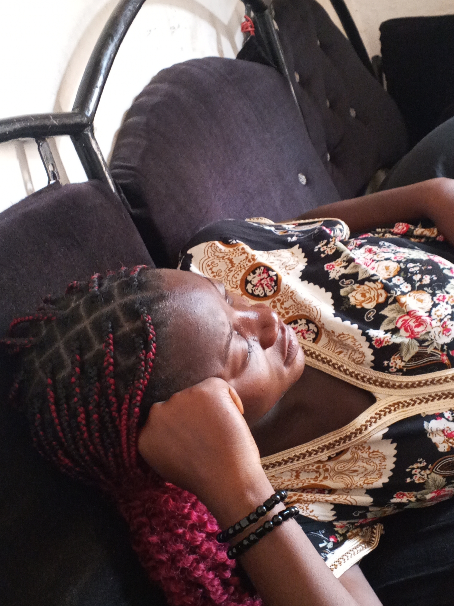 A woman lies on a dark-colored sofa in an indoor setting, potentially a home. She is wearing a floral-patterned dress with cream-colored detailing. Her head rests on a hand covered in red braided hair. She has two beaded bracelets on the arm supporting her head. The sofa is upholstered with buttoned cushions and has a black metal frame. The location is unknown and not in the city of Gwong, Nigeria.