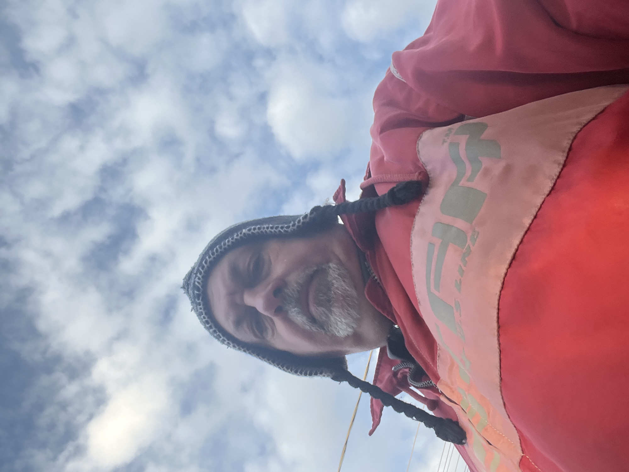 A low-angle shot shows a person wearing a knitted hat with braided extensions and a red jacket, against a background of a cloudy sky. The person has a gray beard and is looking down at the camera. The jacket appears to have a light pink panel with possibly lettering or a logo. The location in Podolsk, Russia, is not discernible from the image.