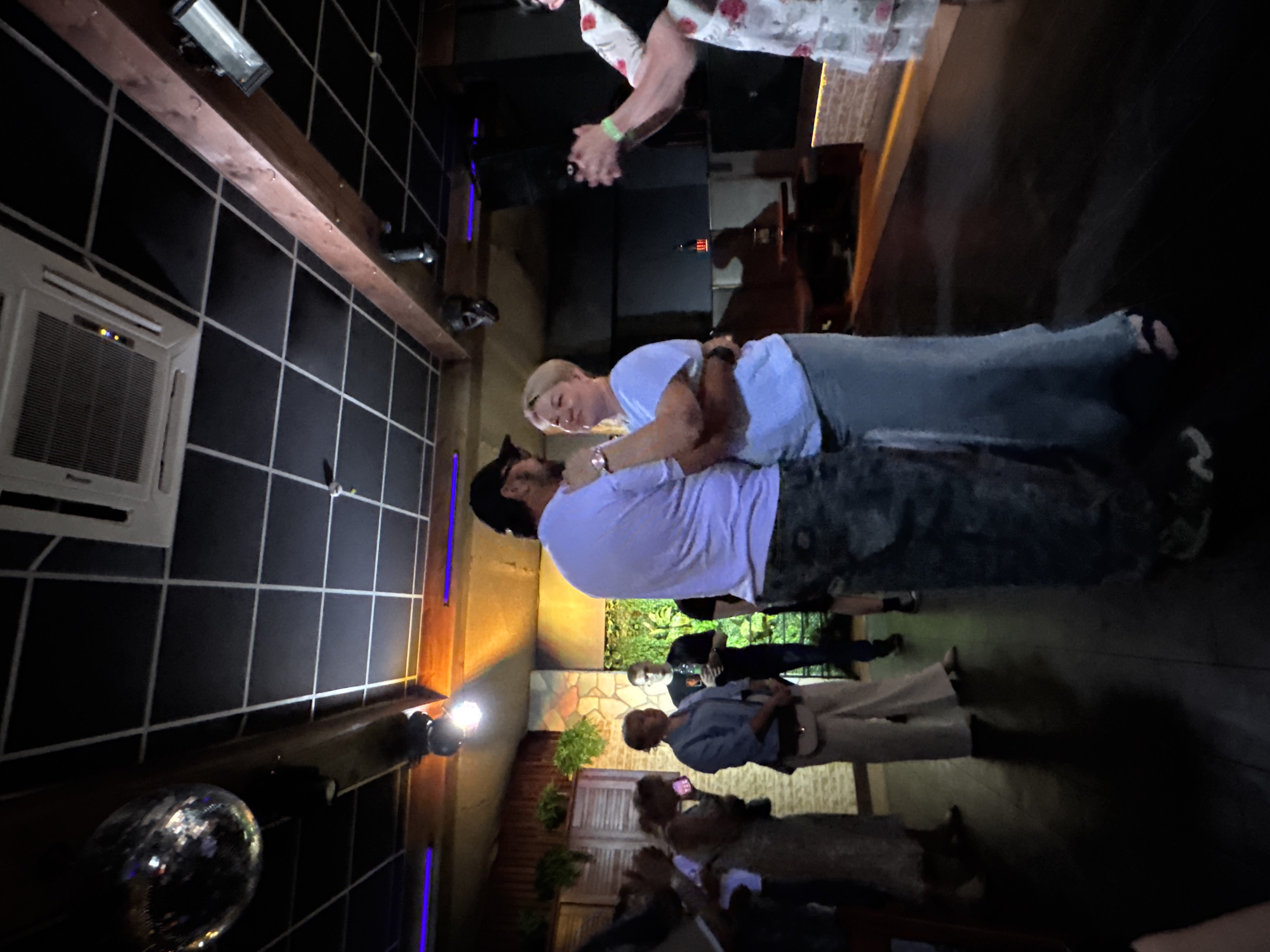 The image shows the interior of what appears to be a nightclub or lounge, likely in the city of Chaikovsky, Russia, although this cannot be definitively confirmed from the image alone. Two people are dancing closely in the center of the frame. A woman with short blonde hair is wearing a light blue shirt and jeans, while a man with a beard and a black cap is wearing a light purple shirt and camo pants.
A crowd of people, blurred and indistinct, are observed near a stone wall or partition. Some are interacting and possibly filming or photographing the dancers. One individual is seen with a camera, possibly taking photos. The interior features dark walls tiled in a grid pattern, with some lighting fixtures attached to the ceiling and walls. A disco ball is visible in the lower left corner.