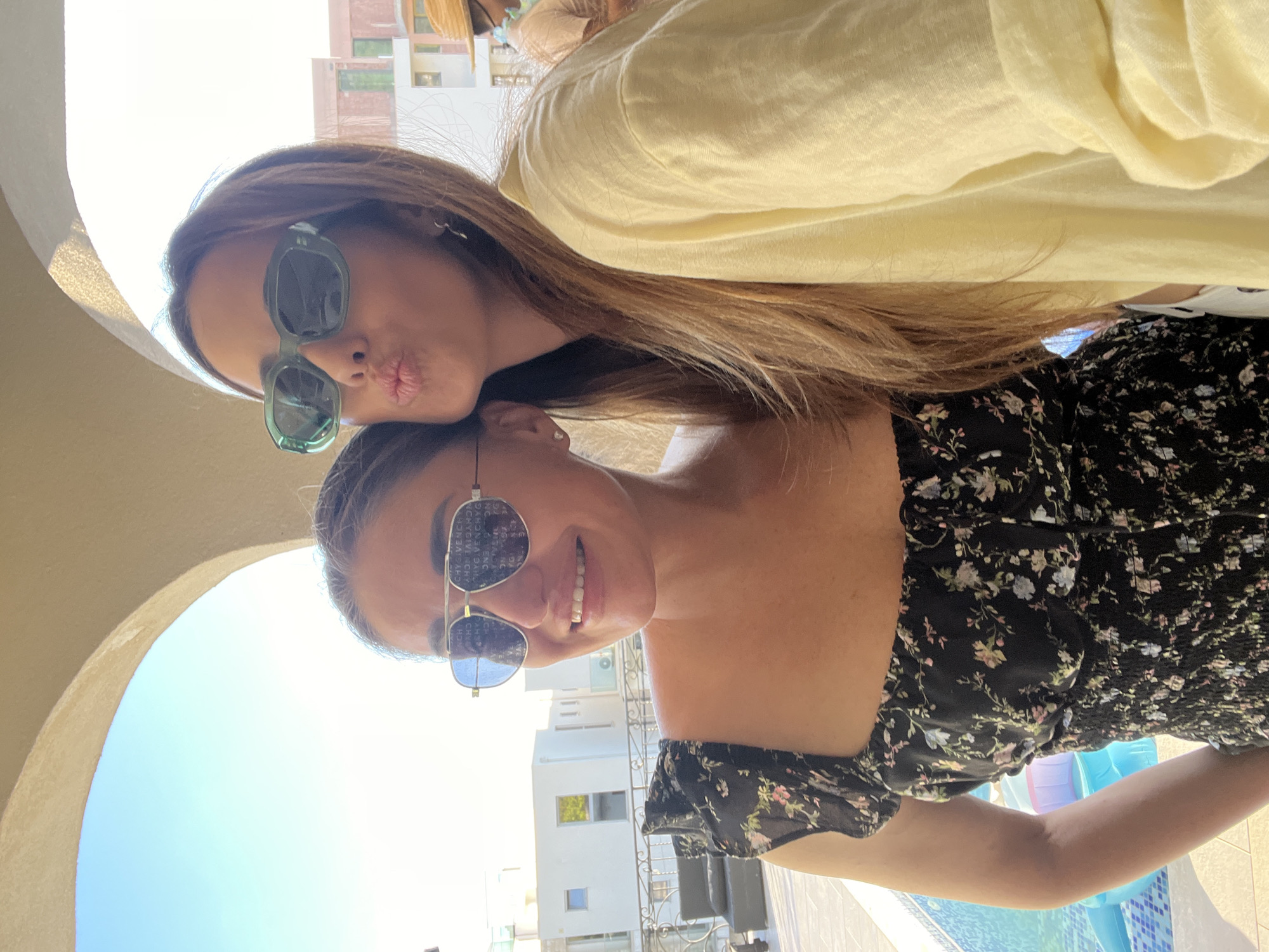 Two women are posing for a selfie outdoors. One woman, wearing a black floral dress and silver-framed sunglasses, is smiling. The other woman, wearing green-framed sunglasses and a yellow top, is puckering her lips as if to kiss the other woman. A glimpse of a white building, likely a hotel or resort, is visible in the background. A sliver of a pool with blue inflatable pool toys is visible on the right. An arched structure is partially visible in the background, possibly a part of the building or a surrounding structure. The location is likely Dobra Voda, Montenegro, given the white architecture and pool environment.