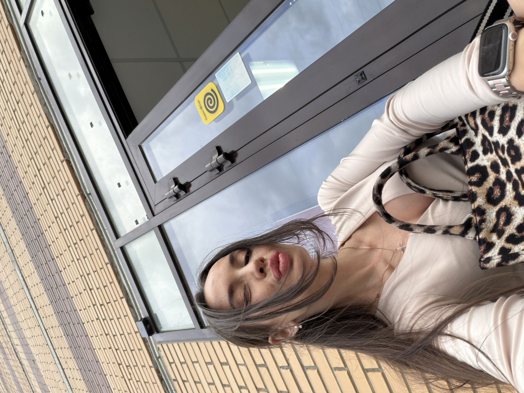A woman with long, dark brown hair and fair skin is captured in a close-up, low-angle selfie. She winks with her right eye, and her lips are pursed, facing slightly upwards. She wears a light beige, long-sleeved V-neck top and a thin gold chain necklace with a small cross pendant. A small gold hoop earring with a clear stone is visible in her left ear. On her left wrist, she wears a silver-toned smartwatch with a metallic link band. She holds a leopard-print handbag with her right hand. The background features a brick building exterior with alternating light beige and darker brown horizontal brick bands. Below the brickwork is a dark-framed glass door or window. Affixed to the glass are a yellow square sticker with a black spiral logo and the Cyrillic text "Яндекс.Еда" (Yandex.Food), and a smaller, light blue rectangular sticker. Two dark metal hinges are visible on the door frame. Above the door, a dark rectangular object is mounted on the brick wall, connected to a metal conduit. The reflections in the glass indicate an overcast sky. The "Яндекс.Еда" sticker identifies the location as Russia, specifically within the city of Saint Petersburg where the service operates.