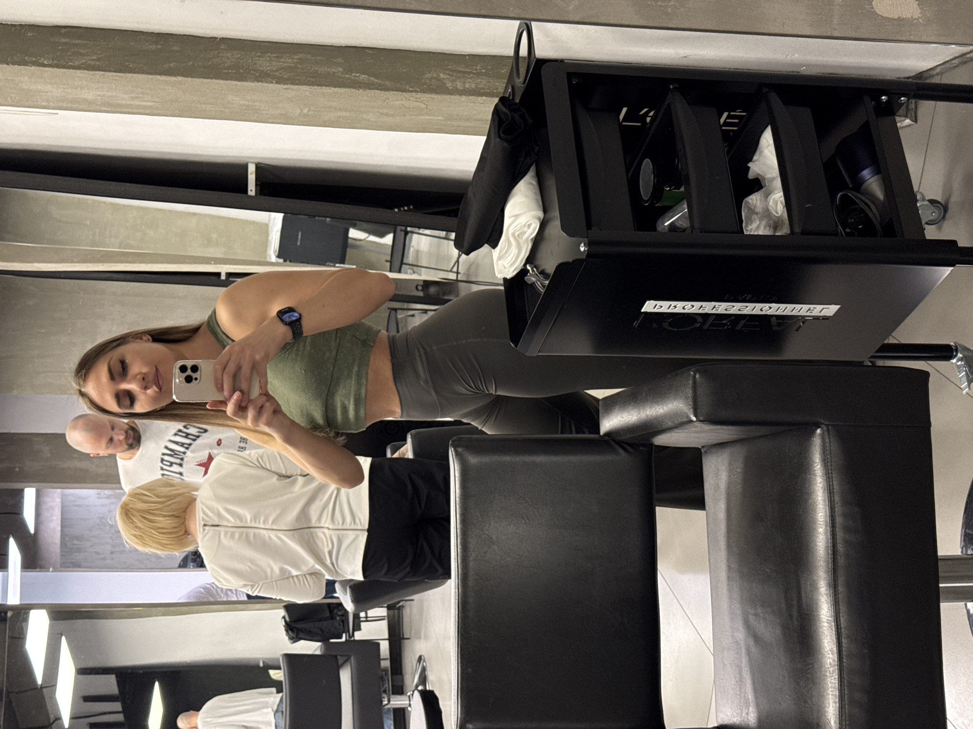 A selfie in what appears to be a hair salon. A woman with long brown hair and wearing a green athletic top and gray pants is holding a smartphone and taking a photo. Behind her to her left is a man with a shaved head wearing a white t-shirt. In front of her to the right is a woman with blonde hair wearing a white jacket. In front of them are black chairs and a black rolling cart with hair styling supplies. The salon has grey walls and mirrored surfaces. There is no information to indicate the location is Saint Petersburg, Russia.