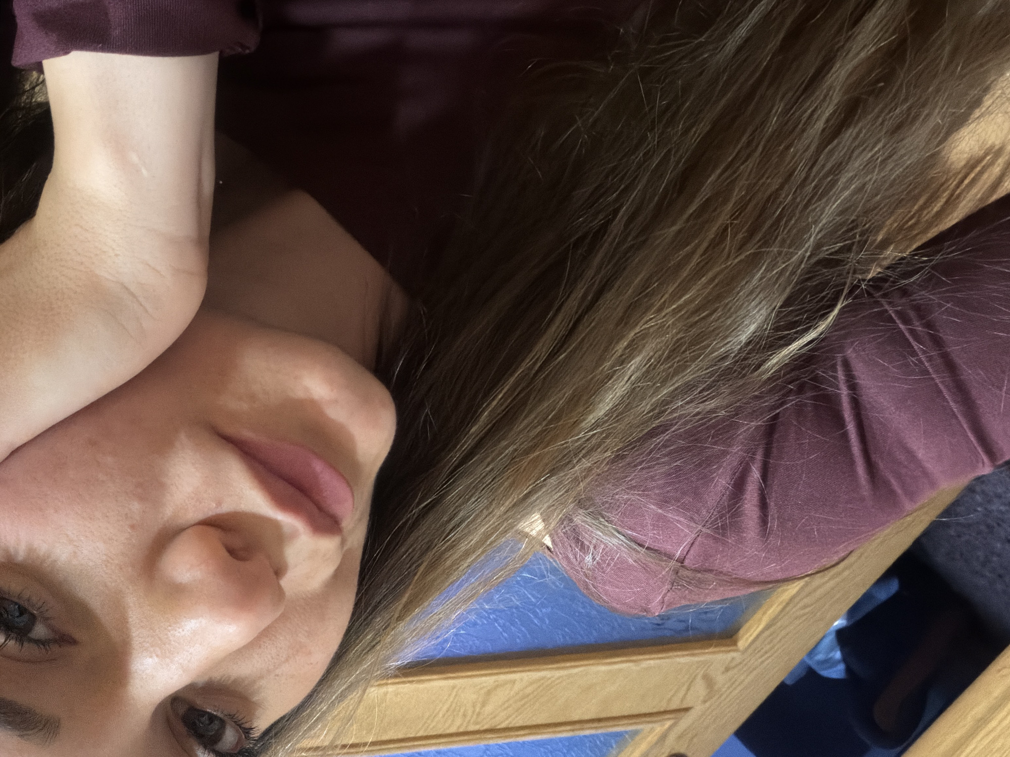A medium close-up image of a person with light brown hair, and a maroon shirt. The person is resting their head on their hand. In the background is blue and brown wood, and a dark gray fabric. No location can be confirmed.