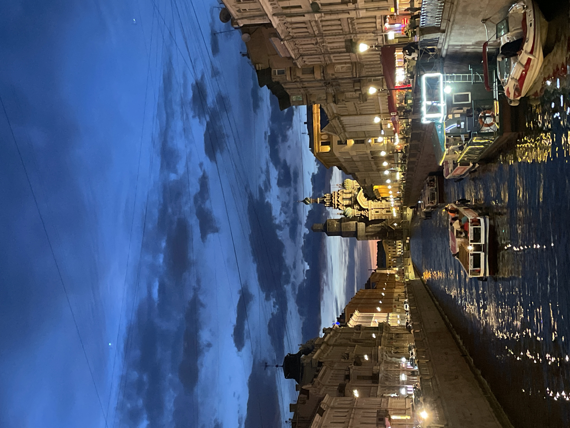 A vibrant twilight or early night scene captures a bustling canal in Saint Petersburg, Russia, flanked by illuminated historic architecture. The sky is a deep, rich blue with scattered, dark clouds, characteristic of the "blue hour," and numerous utility wires stretch across it.
The main focus is the tranquil canal, where several small passenger boats navigate the waterways. These boats, some brightly lit, carry indistinct figures, suggesting tourists or locals enjoying an evening cruise. The water's surface reflects the myriad of artificial lights from the buildings and boats, creating a shimmering, dynamic pattern.
Along both banks of the canal, grand, multi-story buildings, adorned with classical architectural details, are brightly illuminated, showcasing their intricate facades. Streetlights line the embankments, casting warm glows on the pathways and contributing to the radiant reflections in the water.
In the background, a distinctive landmark rises above the cityscape: a prominent, multi-domed church, clearly identifiable as the Church of the Savior on Spilled Blood, its unique spires and ornate details glowing under the night lights. This iconic structure firmly places the scene in Saint Petersburg.
The overall atmosphere is one of serene urban beauty and historical grandeur, highlighting the city's famous waterways and architectural heritage under the enchanting light of dusk. No discernible text is visible in the image.