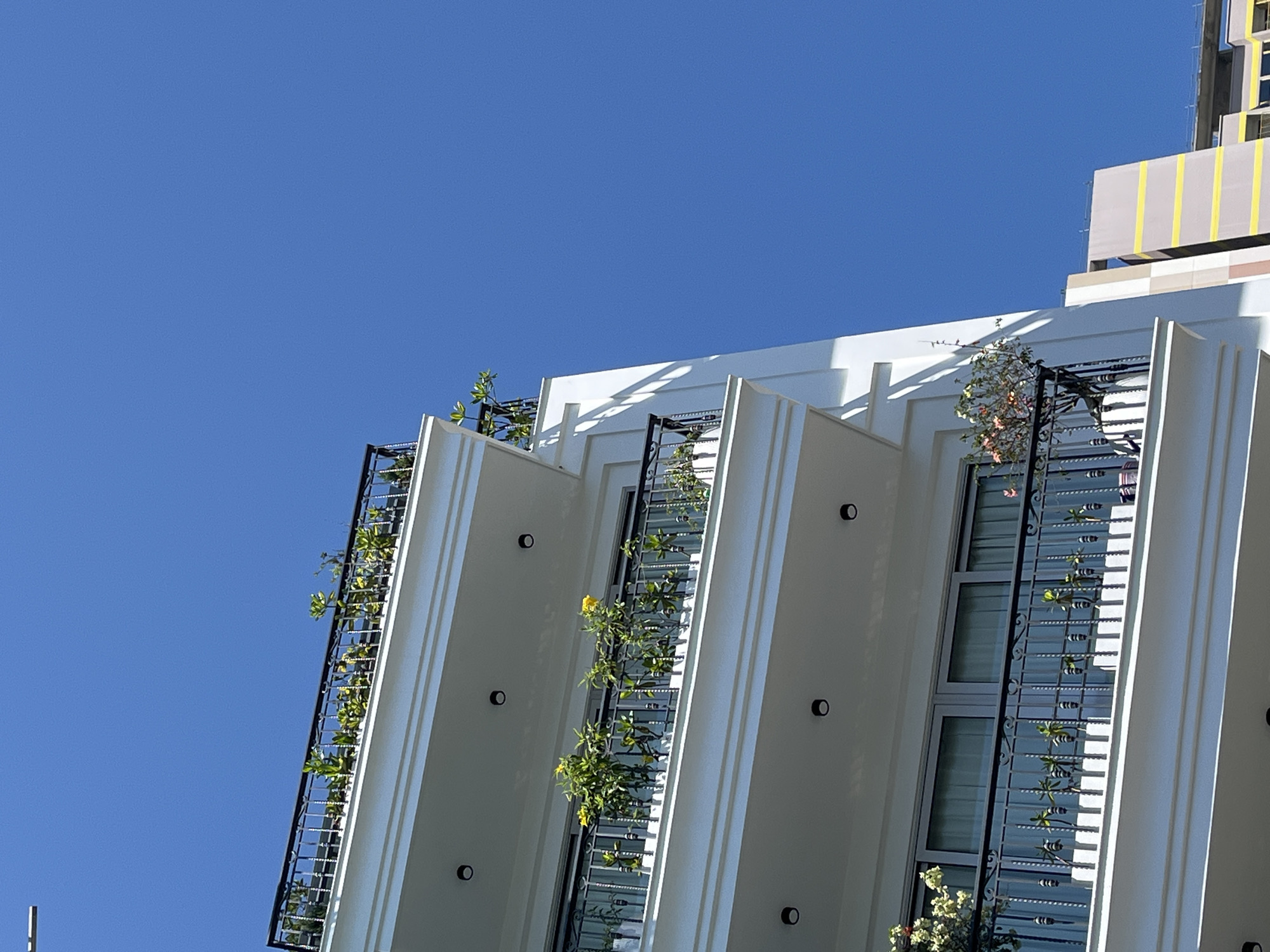 The image shows a low-angle view of a building, likely an apartment complex, in Đà Nẵng, Vietnam. The architectural style features white facade with vertical protruding sections, each fitted with black metal railings and planters containing greenery. The building has a modern aesthetic, with clean lines and cylindrical external lights on the white walls. A taller structure, possibly another building under construction, is visible in the background, identifiable by its yellow and grey tarp. The sky is a clear, vibrant blue.