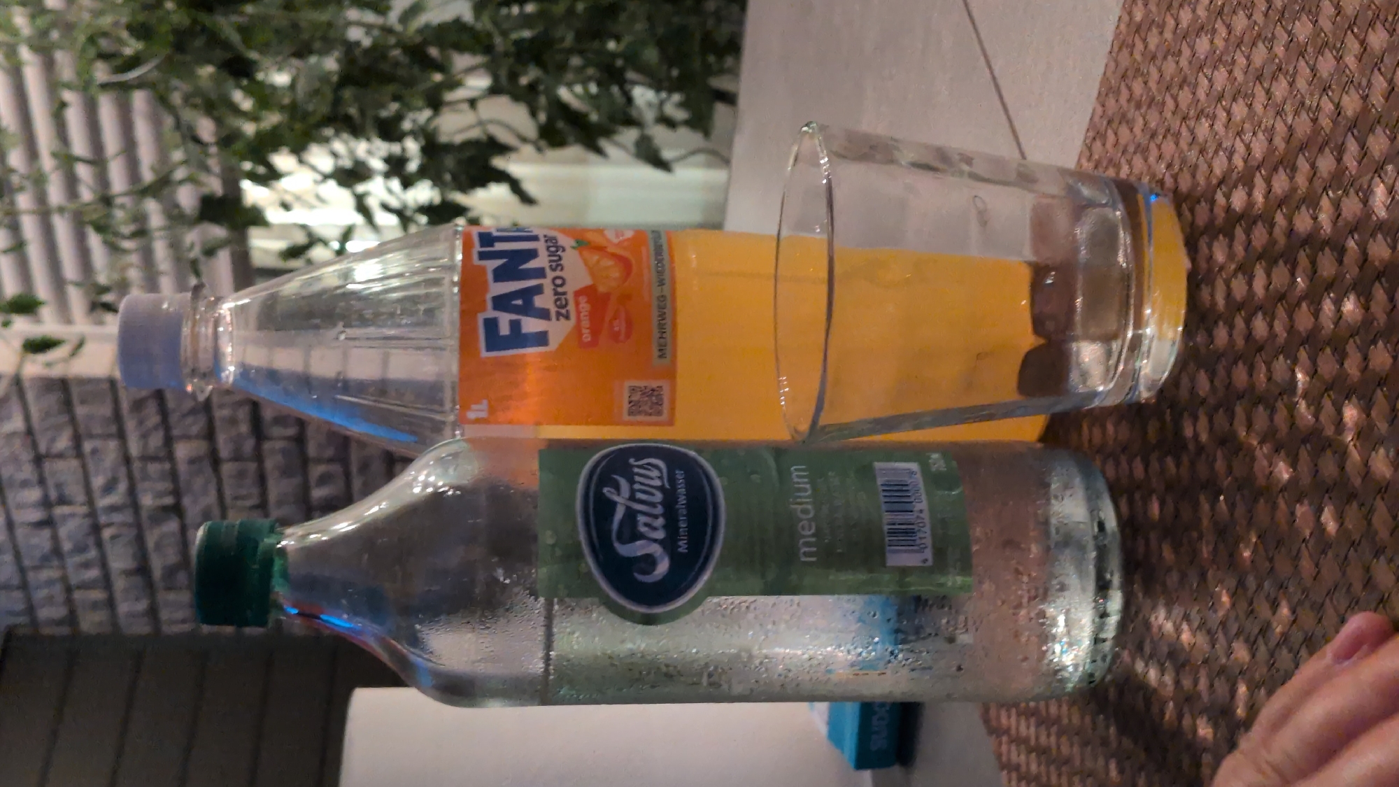 The image displays two plastic bottles and a glass on a textured surface, likely a table with a woven placemat. The bottle in the foreground is filled with clear water and has a green cap. Its label is green and white and reads "Salvus Mineralwasser medium". The bottle behind it contains an orange beverage and has an orange and white label that says "FANTA zero sugar orange". Beside the Fanta bottle is a clear glass filled with the same orange liquid. The scene appears to be outdoors, possibly a patio or garden area, with lush green foliage visible in the background. The lighting suggests it could be daytime. No people are clearly visible in the image, although a hand is partially visible in the lower right corner. The overall setting evokes a casual, relaxed atmosphere, perhaps a break or a refreshment stop.