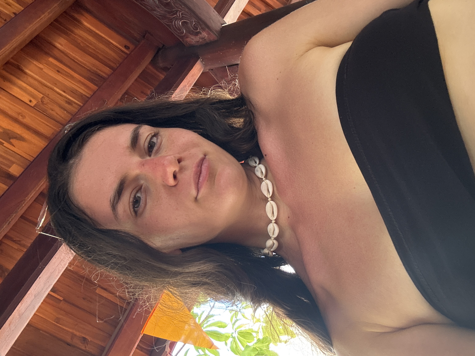 A young woman is looking into the camera, wearing a black top and a necklace of seashells. She is under a wooden structure with a tiled roof. Vegetation is visible in the background, suggesting an outdoor location. Based on the prompt mentioning Mũi Né, Vietnam, the scene likely takes place in a resort or beach setting in that city.