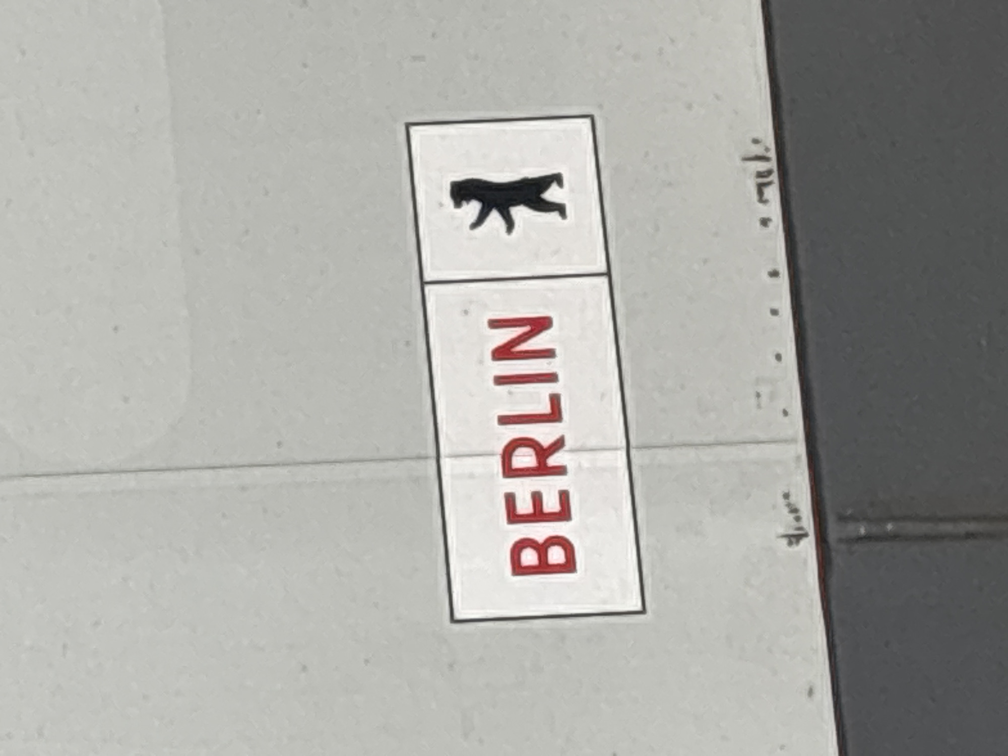 A rectangular white sticker, outlined with a thin black border, is affixed to a textured, light gray vertical surface. On the left side of the sticker, the word "BERLIN" is displayed in red capital letters. The right side features a black silhouette of a bear standing on its hind legs, facing right, with its front paws slightly raised. The light gray background surface exhibits subtle vertical and horizontal seams, along with scattered small, faint dark specks. Immediately beneath the sticker, on the light gray surface, there are minute, indistinct dark markings. A narrow red horizontal line separates the light gray surface from a dark gray horizontal panel below. The scene is located in the city of Schönefeld, Germany.