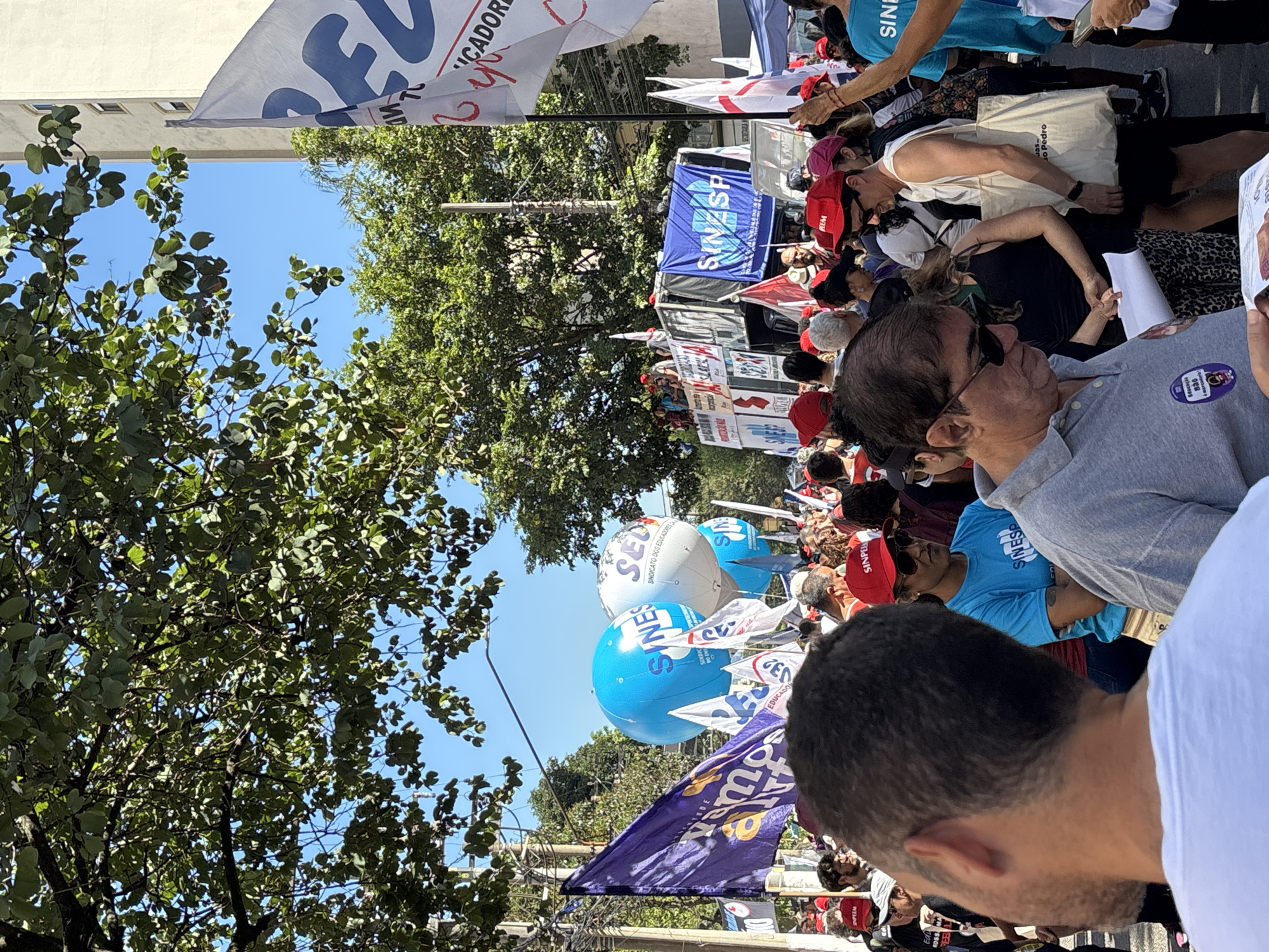 A crowd of people are visible, seemingly participating in a march or demonstration, likely in the city of São Paulo, Brazil. Many individuals are carrying flags and banners associated with various unions and educational organizations, including SINESP, SINPEEM, and SED. There are several blue and white inflatable balloons bearing the SINESP logo. People are dressed casually, and red hats with "SINPEEM" printed on them are visible. The surroundings consist of urban scenery with trees and buildings.