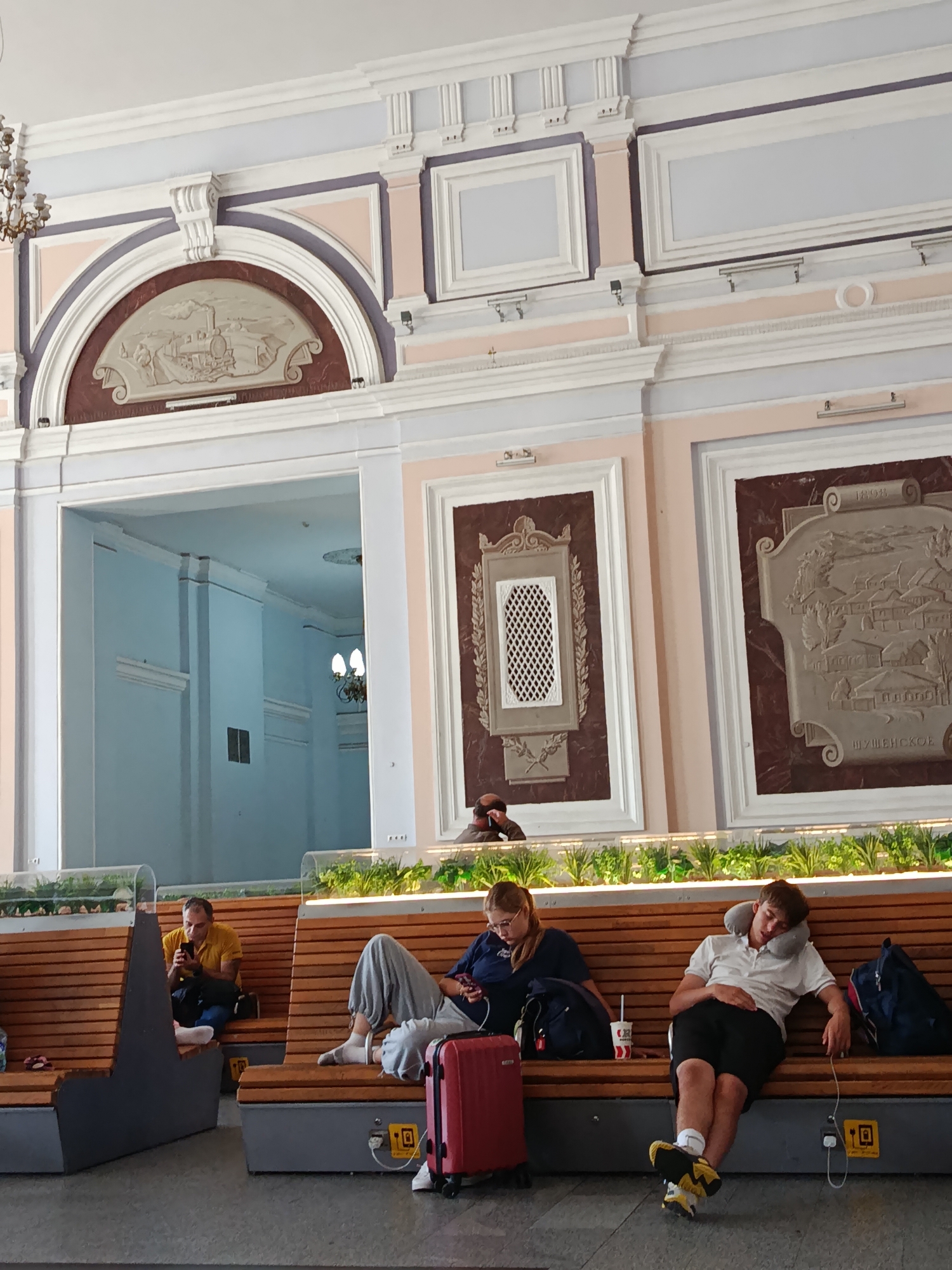 The image depicts the interior of a transportation hub, likely a train station, given the presence of seating areas and the overall architectural style. The location is identified as Moscow, Russia. The scene is brightly lit, suggesting it's daytime.
In the foreground, several people are seated on wooden benches. On the left, a man in a yellow shirt is looking at his phone. Next to him, a woman with glasses is also engaged with her phone, with a red suitcase beside her. On the right, a young man is asleep on the bench, with a neck pillow supporting his head and a backpack on the floor next to him. Another person, partially visible, is seated to his right.
The benches are arranged in rows, with a partition of greenery separating them from a backdrop of ornate wall decorations. These decorations include framed reliefs and architectural moldings, characteristic of older public buildings. The walls are painted in pastel colors, with a light pink and a light blue predominating. In the background, a large arched opening reveals a hallway or another section of the building, also featuring decorative elements and lighting fixtures. The floor appears to be tiled. The overall impression is one of a well-maintained, albeit somewhat dated, public space designed for travelers.