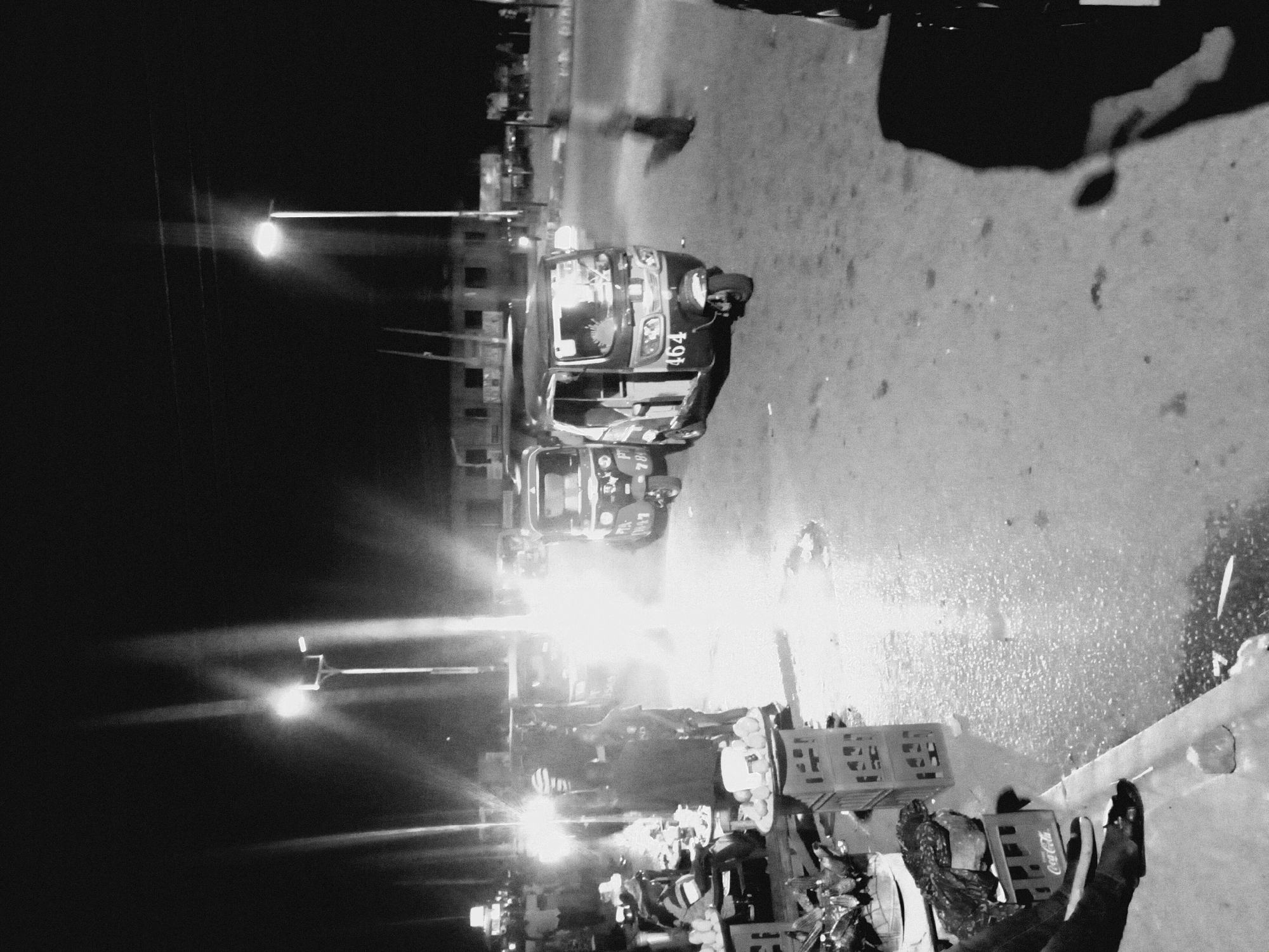At night in Bukuru, Nigeria, a black and white image shows a street scene with bright streetlights and vehicle headlights. An auto-rickshaw marked with "464" is visible. There are people, objects, and Coca-Cola crates in the foreground. The street appears wet, reflecting light.