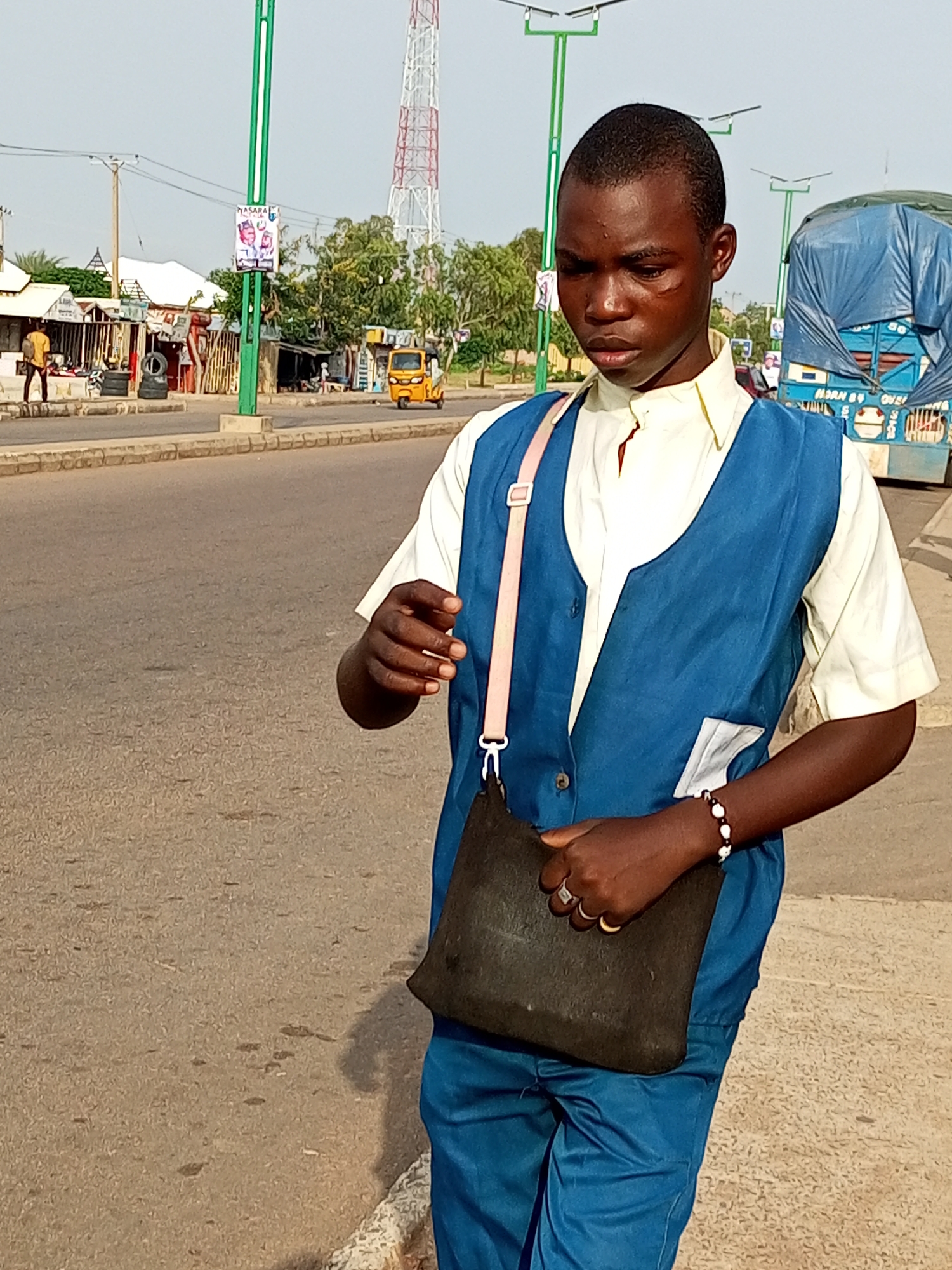In Tumfure, Nigeria, a young man stands on a paved roadside, looking down as he holds a dark shoulder bag. He is wearing a light-colored collared shirt and a blue vest with a light pink strap from the bag crossing his chest. A beaded bracelet adorns his left wrist. The background shows a street with shops, a yellow auto-rickshaw, a large blue truck covered with a tarp, and a radio tower in the distance. The sky is clear and the scene appears to be illuminated by natural daylight.