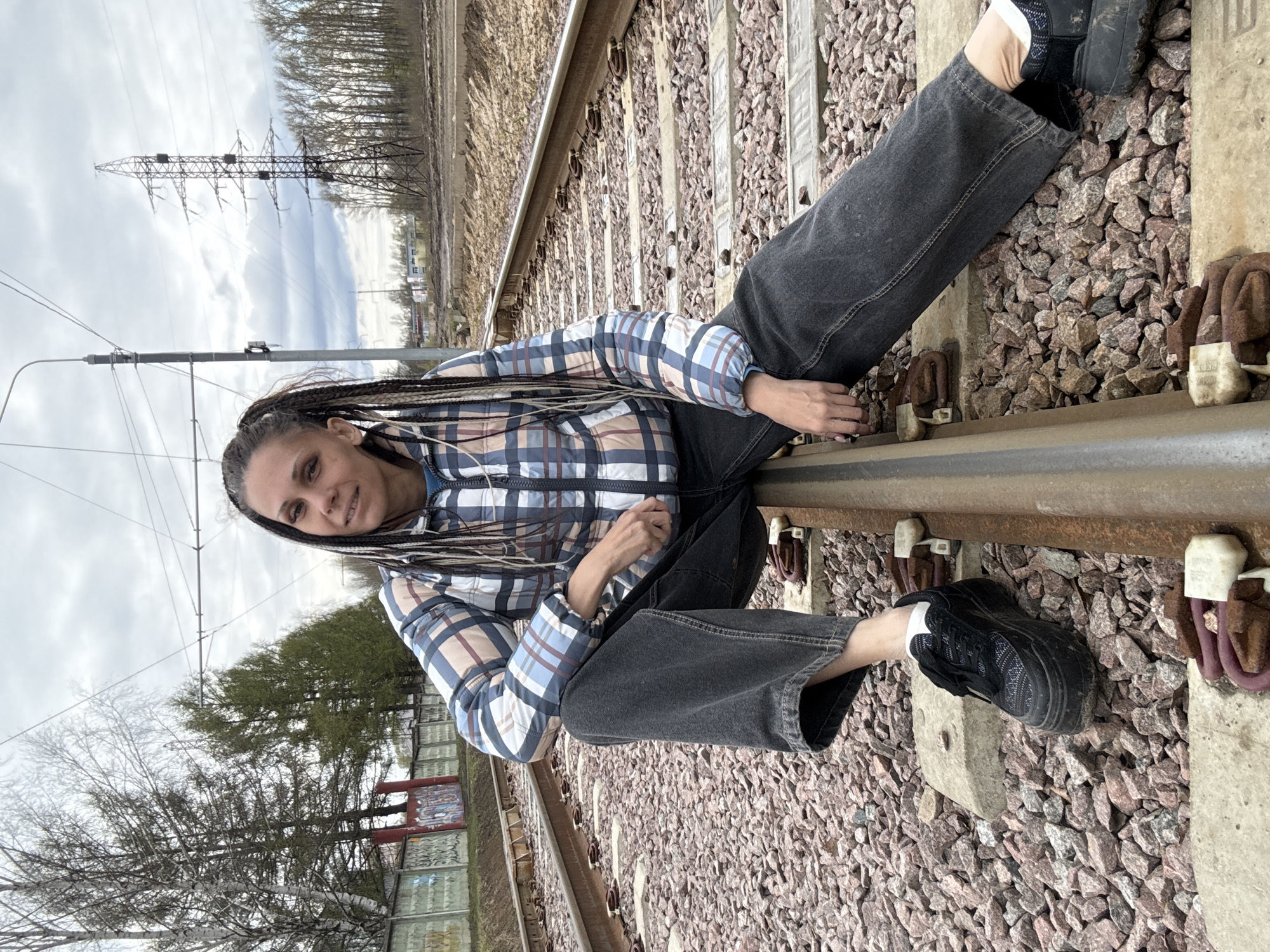 This image, taken in Заволжье, Russia, depicts a woman sitting casually on railway tracks in an outdoor setting. The photograph is oriented vertically, with the woman positioned diagonally across the frame.
The main subject is a woman with long, dark braided hair, which includes lighter highlights or extensions. She is wearing a blue, brown, and white plaid puffer jacket or vest over a blue top, paired with dark, loose-fitting jeans and black athletic shoes with visible white socks. She is looking directly at the viewer with a calm, slight smile. Her posture is relaxed, with her legs extended along the tracks, one foot on the gravel ballast and the other on a concrete sleeper. Her hands rest on her lap or near the rail.
The setting is a railway line, featuring metal rails, concrete sleepers, and a bed of gravel ballast. Fasteners securing the rails to the sleepers are visible. The background reveals an open landscape under an overcast sky. A prominent metal lattice power line tower stands on the left side of the background. Beyond the tracks and tower, a line of bare trees suggests either late autumn or early spring. Another utility pole is visible further back, and indistinct structures or fencing with some markings can be seen in the distance to the right.
The weather is cloudy with no direct sunlight, indicating an overcast daytime. Visible text on the concrete sleepers includes "ЦП 34", "Б1", and numbers such as "10" and "1410" on rail fasteners, likely identification codes.