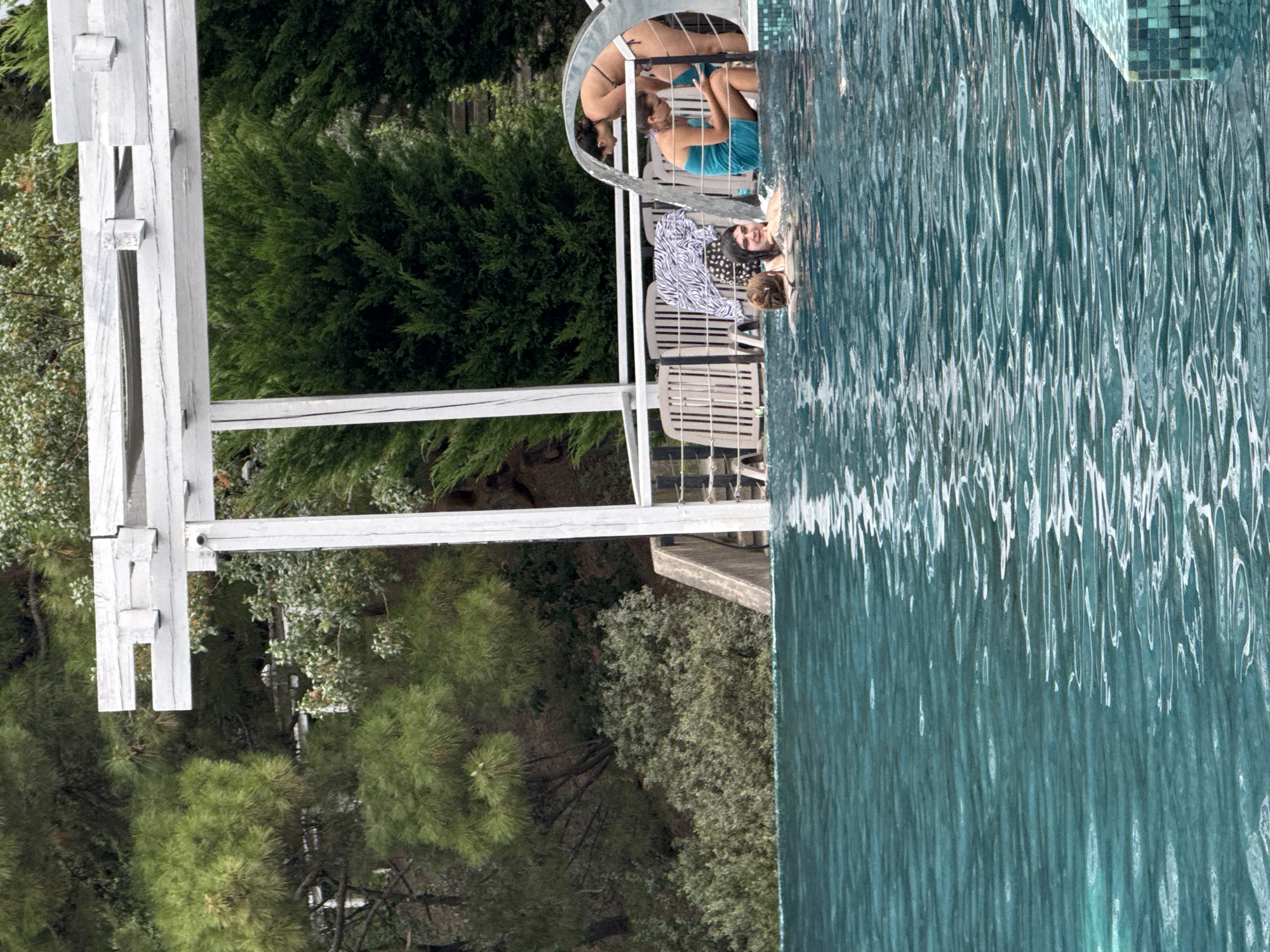A group of individuals are in a swimming pool located in a lush, green outdoor environment. The pool is filled with turquoise water and appears to be an infinity pool, with the edge of the pool and the tiled border visible. In the background, there is a white pergola structure supported by white posts. Behind the pergola, there are chairs and a patterned blanket. The area is surrounded by dense green foliage, including trees and bushes, suggesting a natural setting. There are at least four people visible in the pool. One person in a blue swimsuit is partially submerged, facing away from the camera. Another person in a bikini is bending over, appearing to interact with someone in the blue swimsuit. Two more individuals are visible in the water, with only their heads and shoulders showing. The overall scene depicts a relaxed outdoor gathering by a pool.