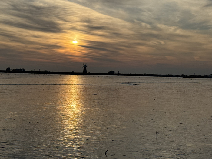 The image presents a panoramic view across a wide body of water, identified as the River Yare in the Great Yarmouth area, United Kingdom, at either sunrise or sunset.
The sky, occupying the upper third, features layered clouds ranging from dark grey-blue at the top to vivid orange and yellow around the prominent, circular sun positioned slightly left of the image's vertical center. An elongated, intense golden-orange reflection of the sun stretches across the calm water surface, dominating the central and lower portions of the frame.
The horizon line, located approximately one-third from the bottom, is a dark silhouette of land. From left to right, it displays a cluster of indistinct buildings, several vertical posts, the distinct four-sailed silhouette of Berney Arms Windmill, and a lone tree. Small, dark specks are present on the distant horizon and in the upper sky, consistent with birds.
The water, occupying the lower two-thirds, is generally calm with minor surface ripples, reflecting the sky's muted tones of dark grey and brown, intensely illuminated by the sun's reflection. A faint, dark, arcing line is visible on the water's surface in the mid-ground, left of the main reflection. Two thin, dark vertical sticks protrude from the water in the lower right quadrant, and scattered debris appears on the water's surface. No human subjects are visible.