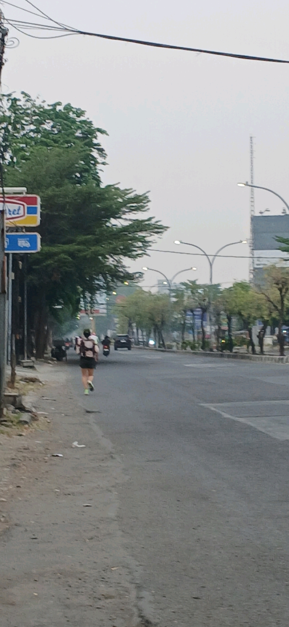 The image depicts a street scene in Cirebon, Indonesia, likely during early morning or late afternoon due to the diffused lighting. On the left side of the road, a person is running on the sidewalk. The runner is wearing a dark athletic outfit with a patterned backpack. Further down the sidewalk, there is a street lamp and a sign for "Indomaret," a convenience store chain prevalent in Indonesia. The road itself is paved and appears to be a main thoroughfare, with several vehicles visible, including a black SUV and motorcycles. Trees line both sides of the road, and street lamps with curved arms are present. The sky is overcast and hazy. The general atmosphere suggests an urban setting in Indonesia, with a focus on the activity of the runner against the backdrop of city infrastructure and traffic.