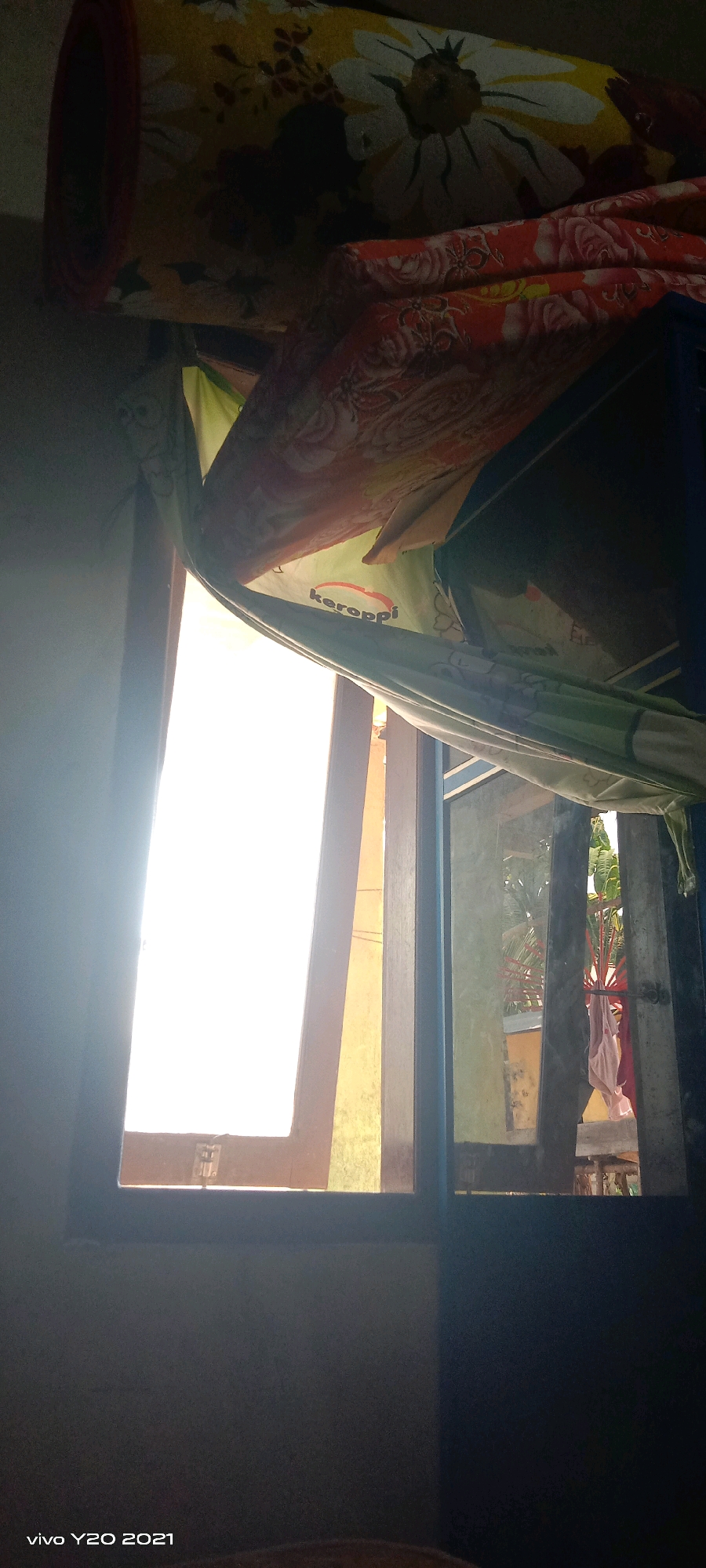 The image shows a close-up of an open window with bright sunlight streaming in. A floral patterned curtain is partially covering the window. In the background, through the window, there are some structures and vegetation. The overall setting appears to be indoors, looking out. The bottom left corner of the image displays "vivo Y20 2021".