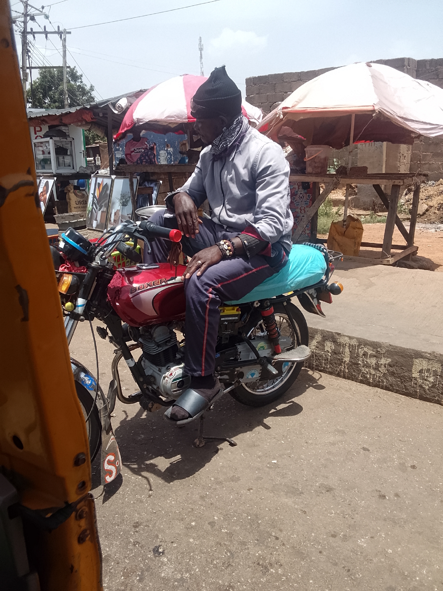 The image depicts a man seated on a red "Boxer" motorcycle on a paved street in what appears to be a commercial area. The man is wearing a dark-colored knit hat, a light gray long-sleeved shirt, dark pants, and slides over socks. He has a patterned face covering pulled down around his neck. He is wearing a watch and wristbands on his left wrist and a ring on his left hand.
The motorcycle is parked with its kickstand down. The scene includes a glimpse of a yellow auto rickshaw/tuk-tuk on the left. In the background are several roadside stalls covered by white umbrellas with red trim. A few people are visible in the background near the stalls.
The location is identified as the city of Bukuru, Nigeria, based on the presence of the Boxer motorcycle and the characteristics of the commercial setting.
