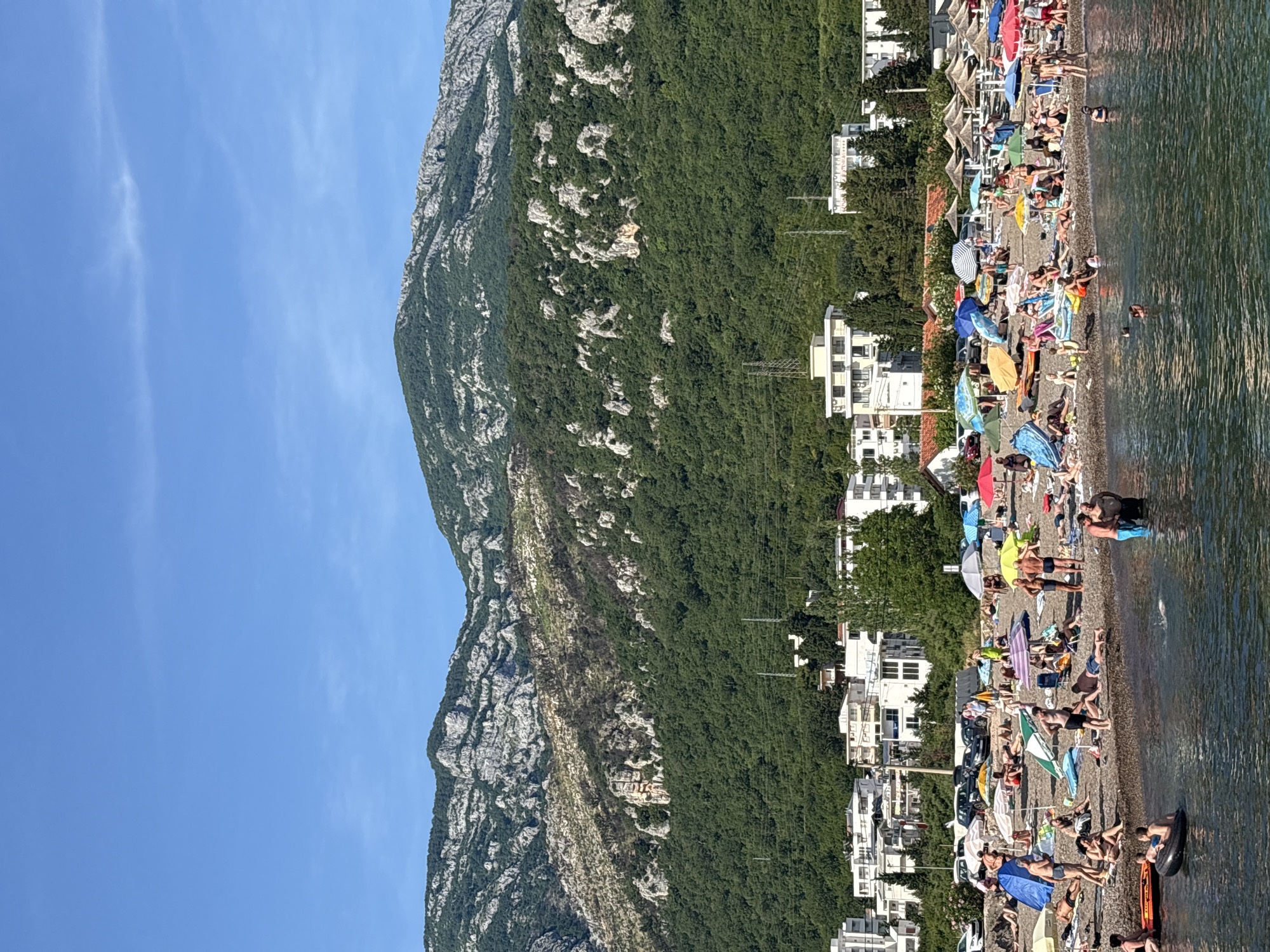 The image depicts a panoramic view of Čanj, Montenegro, on a clear, sunny day. In the foreground, a crowded pebble and dark sand beach extends into calm, greenish-blue seawater. Numerous individuals, primarily adults and some children, are engaged in various activities: sunbathing on towels and sunbeds, sitting under a multitude of colorful parasols (blue, green, red, yellow, striped), walking on the beach, and swimming or wading in the water. One person is observed on a black inflatable ring, and another on an orange inflatable float.
Behind the beach, a row of white and light-colored multi-story buildings with various roof types, including red tiles, indicates a developed coastal town. Several cars are parked on a road or parking area immediately behind these structures. Dense green trees and other vegetation surround the buildings and extend up the slopes of prominent mountains in the background. The mountains feature significant exposed grey rock formations, interspersed with dense green forestation. Patches of drier, lighter-colored vegetation are visible on some lower mountain slopes. The sky above is clear and blue with minimal, faint cirrus clouds. Utility poles and power lines are visible on the lower mountain slopes near the town buildings.