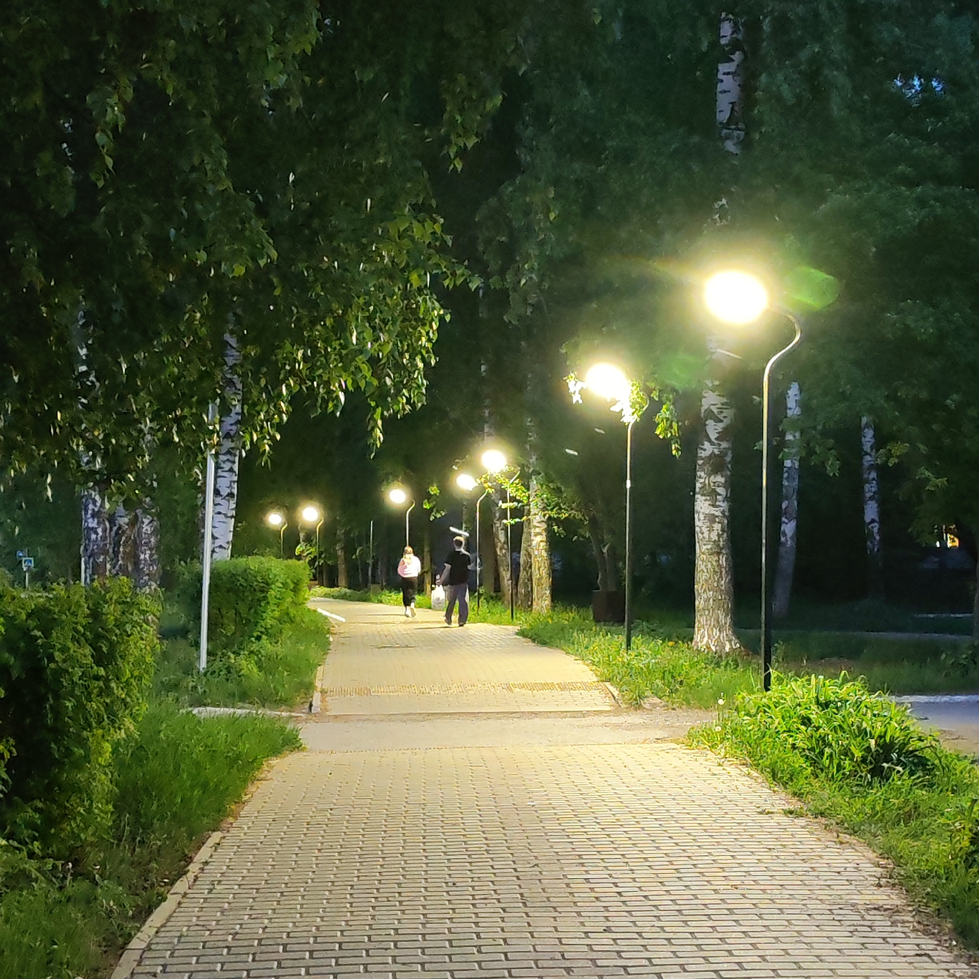 The image captures a brick-paved pathway illuminated by streetlights in a park-like setting, likely in Chaikovsky, Russia. Tall trees with dense foliage line both sides of the path. Two people, a male and a female, are walking along the path in the distance away from the viewer. The male is wearing a black shirt and dark pants, while the female is wearing a white shirt and black pants. The path appears to be well-maintained, with neatly trimmed bushes and grass along the edges. The streetlights cast a bright glow on the scene, with some lens flare effects. The environment suggests a tranquil, possibly residential area within the city. The lighting indicates it is nighttime or late evening.