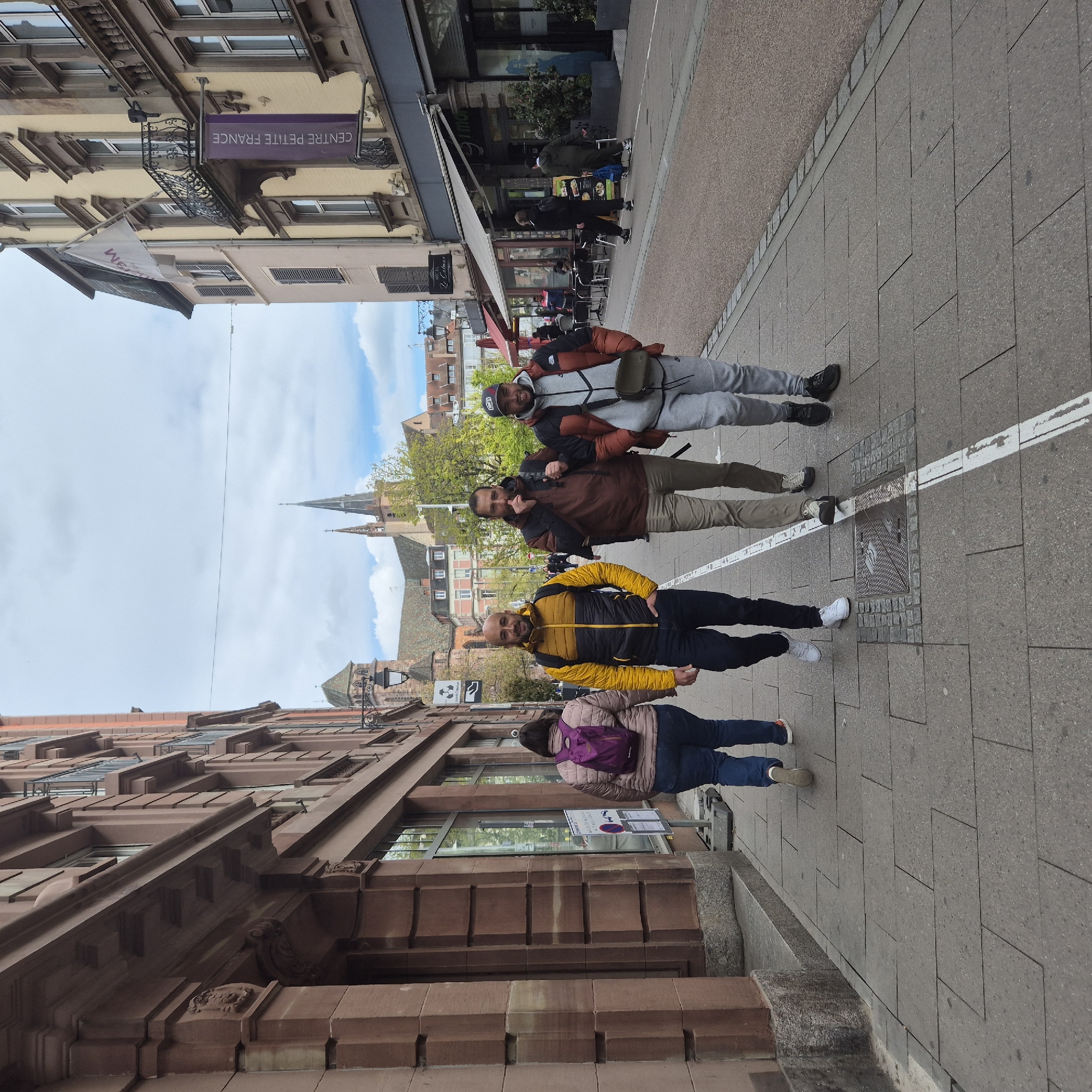 Four people are standing on a sidewalk in Strasbourg, France. The sidewalk is paved with gray rectangular stones. A white line runs along the sidewalk, near a manhole cover.
The first person, closest to the foreground, has dark hair, wears a tan puffer jacket and blue jeans, and carries a purple backpack. The second person wears a yellow and black jacket and black pants with white shoes. The third person wears a brown jacket and tan pants. The fourth person wears a gray tracksuit with a red and black jacket over it, and a black baseball cap.
Behind them are buildings, one with a sign that says "CENTRE PETITE FRANCE." A church steeple is visible in the background. The sky is partly cloudy.
