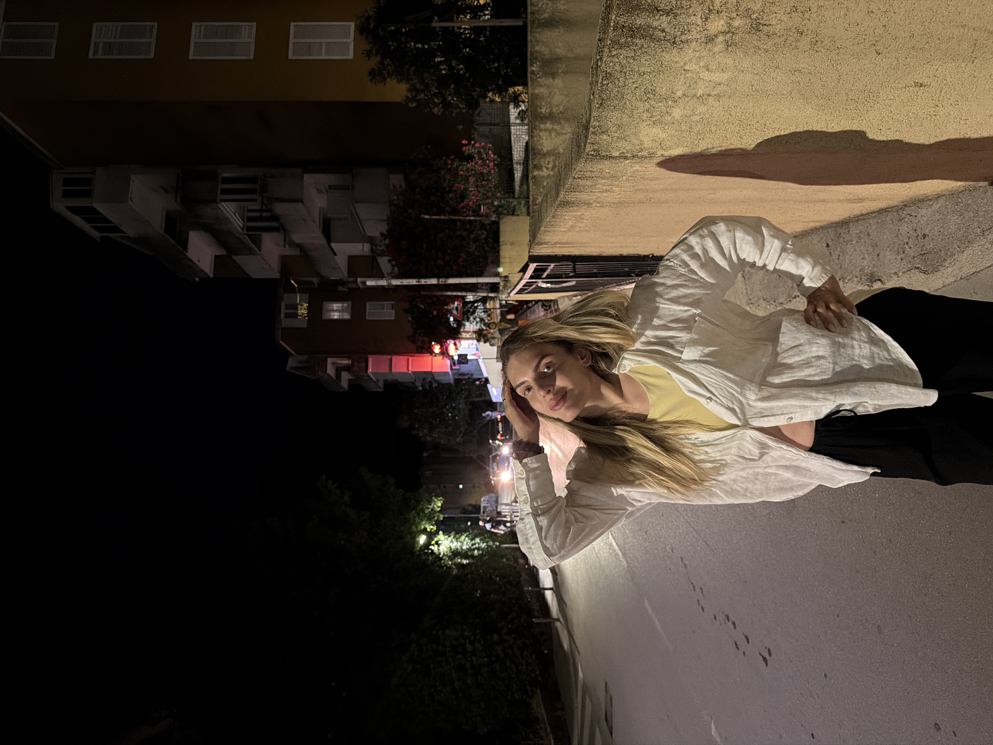 A young woman with blonde hair is standing on a street at night in Budva, Montenegro. She is wearing a white unbuttoned shirt over a yellow top and black pants. Her left arm is raised and her hand is resting on her head, while her right hand is on her hip. She is looking towards the camera with a slight smile. The street is dimly lit, with streetlights casting shadows. In the background, there are buildings, trees, and cars. The overall impression is of a casual evening setting in a European city.