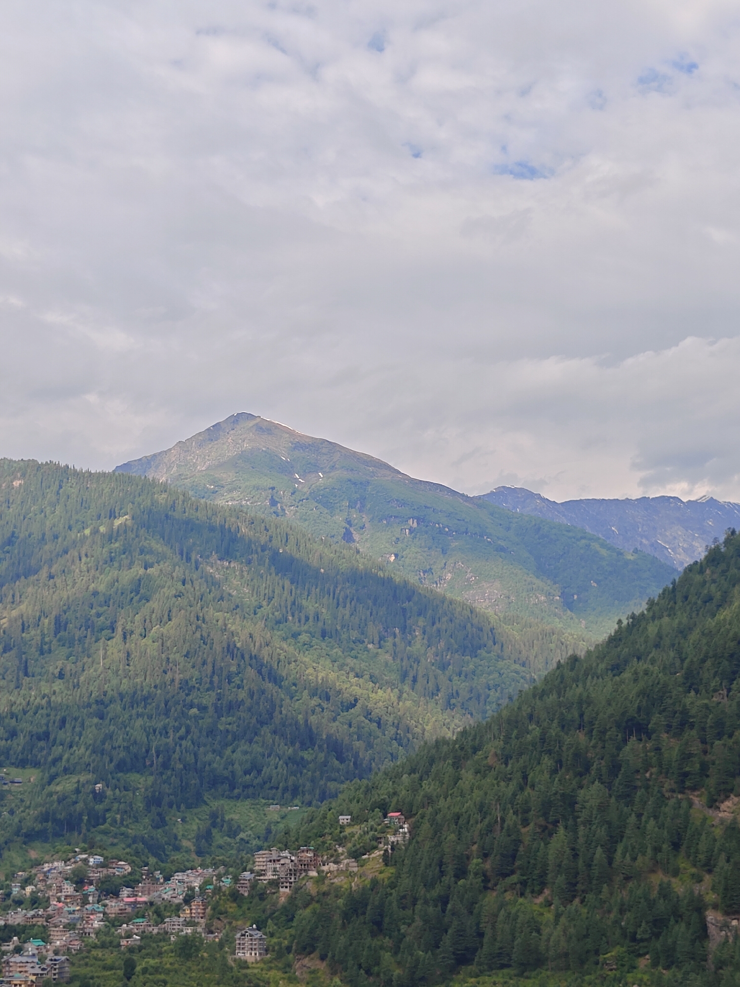 This is a vertical, medium-shot photograph of a mountainous landscape with a town nestled on the slopes. The sky is overcast with layers of grey and white clouds, with patches of blue peeking through. The middle ground is dominated by steep, heavily forested mountains. The trees are a dense, dark green, creating a textured carpet across the slopes. Sunlight casts shadows across the mountains, highlighting the contours of the terrain.
In the foreground, the town of Vashisht is visible on the left side of the frame, built into the side of the mountain. The buildings are a mix of styles, with many appearing to be multi-story structures with flat roofs. Some buildings have balconies and visible windows. The town extends up the hillside, with roads and pathways winding between the houses. Small pockets of vegetation are interspersed with the buildings.
The background features a prominent, sharp peak that is partially snow-capped at its summit. Further back, other mountain ranges are visible, their slopes also covered in vegetation and some showing patches of snow or rock. The overall scene depicts a remote, mountainous area with a human settlement integrated into the natural environment.