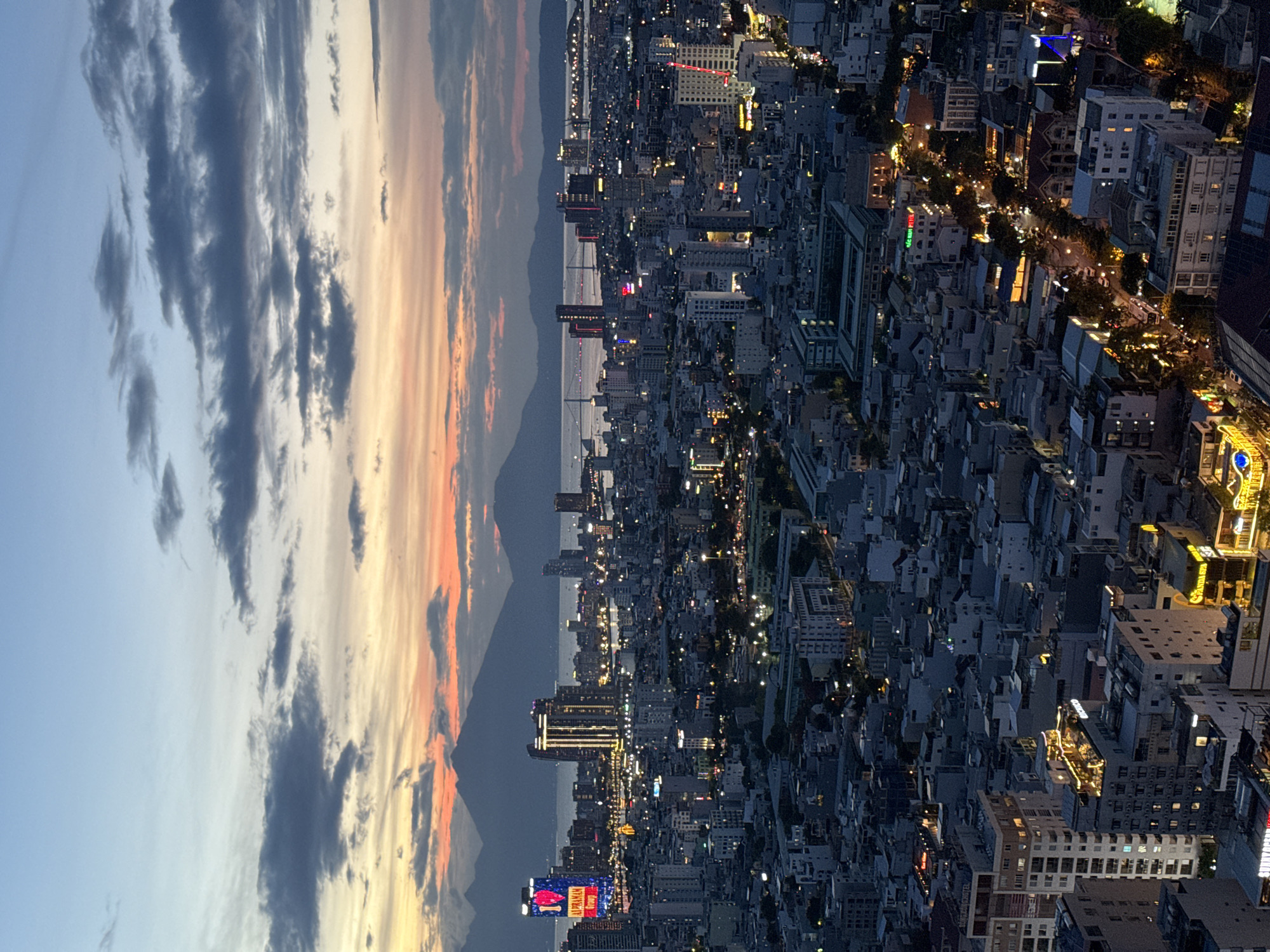 This is a high-angle, panoramic view of Da Nang, Vietnam, captured during sunset or dusk. The image, presented rotated 90 degrees to the left, showcases a vibrant sky transitioning from deep blue on the left to warm oranges, yellows, and pinks on the right, punctuated by scattered, dramatic dark clouds.
Below the colorful sky lies a vast urban landscape. The city is densely packed with buildings of various sizes and architectural styles, extending into the distance. Numerous windows, streetlights, and commercial signs are illuminated, creating a glittering tapestry of lights that signifies the onset of night and the city's activity. In the middle ground, a large body of water, likely the Han River or the sea, can be seen, with several bridges spanning across it, also adorned with lights. Distant mountains are silhouetted against the colorful horizon, adding depth to the scene.
While no individual people are discernible due to the elevated perspective, the illuminated city suggests a bustling environment with ongoing urban life. Notable details include bright red and blue neon signs on some buildings. On a prominent billboard in the lower-left area of the cityscape, visible text includes "LIVE ON" and "SOU" in bright colors. Another yellow sign further right appears to read "PHARMACY" and "HEALTH CARE". The scene evokes a sense of expansive beauty and the energetic rhythm of a city coming alive at night.