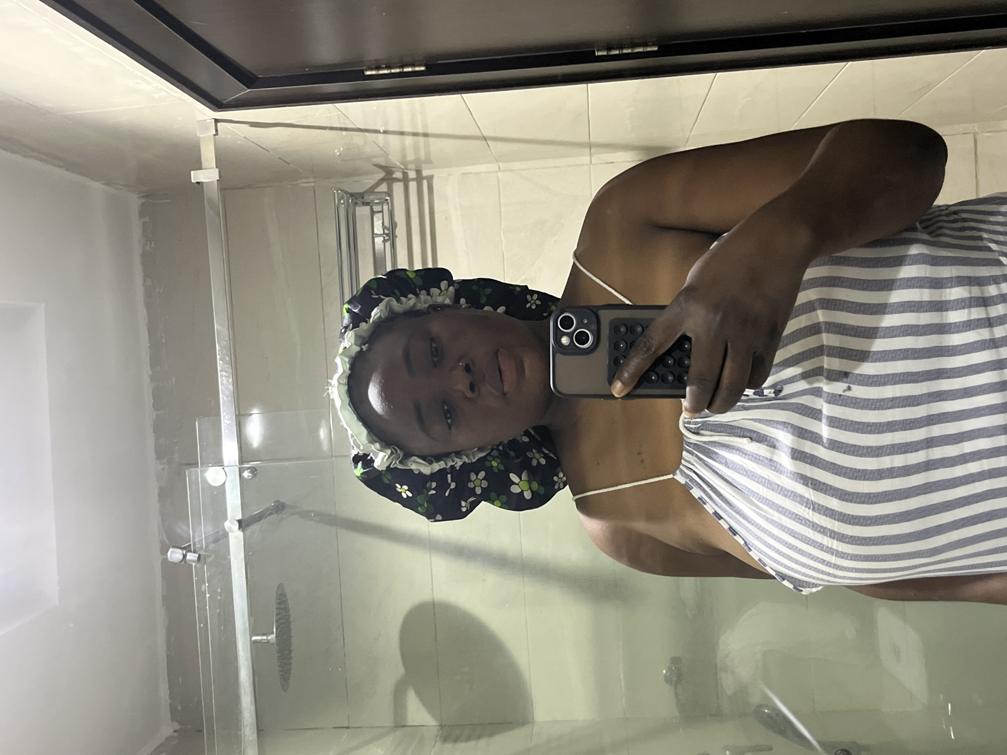 A person wearing a floral bonnet and a striped top is taking a selfie in a bathroom. The bathroom features a shower with a glass door, white tiled walls, and a mirror. It is not possible to infer the location in the city of Abuja, Nigeria, from the image.