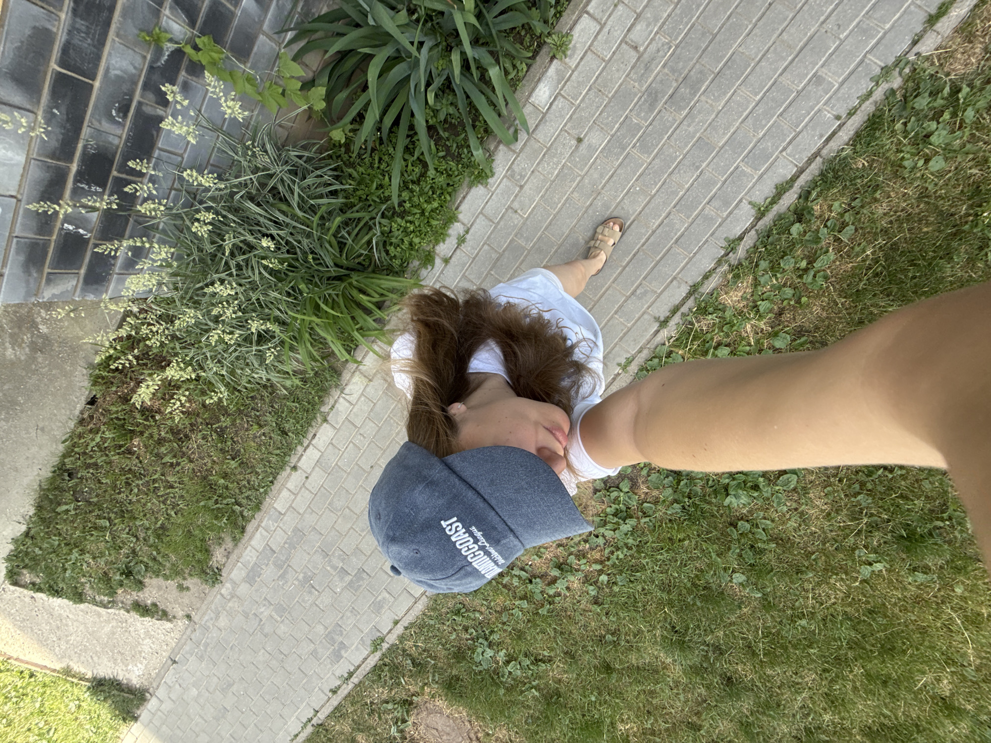 An overhead, high-angle shot shows a person from the chest up, walking on a paved pathway. They are wearing a grey baseball cap with white lettering that reads "Atlantic Coast", a white t-shirt, and beige sandals. Their right arm is extended forward, holding a phone, suggesting a selfie or video recording. The pathway is bordered by a patch of grass on the left and a flowerbed with tall, thin, pale green flowering plants and dark grey brickwork on the right. The scene appears to be outdoors in a residential or public area.
