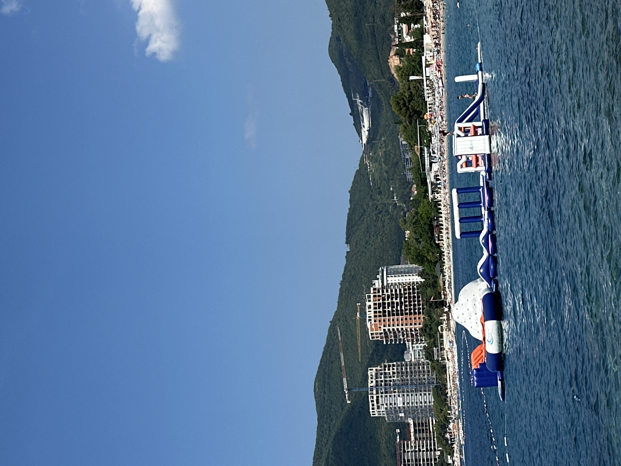 In the city of Boreti, Montenegro, a vibrant scene unfolds on a clear, blue-sky day. The foreground is dominated by the calm, deep blue waters of the Adriatic Sea, where a large, inflatable water park is set up. The park features various interconnected modules, including slides, climbing structures, and platforms, with a prominent white, dome-shaped inflatable. Orange and blue elements are visible throughout the structure. Two individuals are present on the water park, one appearing to be sliding down a ramp, and another standing on a platform.
Beyond the water park, a bustling beach stretches along the coastline. Beach chairs and umbrellas are arranged, indicating a popular recreational area. In the background, several tall, unfinished buildings, presumably under construction, rise against a backdrop of lush, green, hilly terrain. Cranes are visible atop some of these structures, signifying ongoing development. Higher up on the hills, an airplane can be seen in the sky, appearing to be either taking off or landing. The overall environment suggests a lively summer day in a coastal resort town experiencing development.