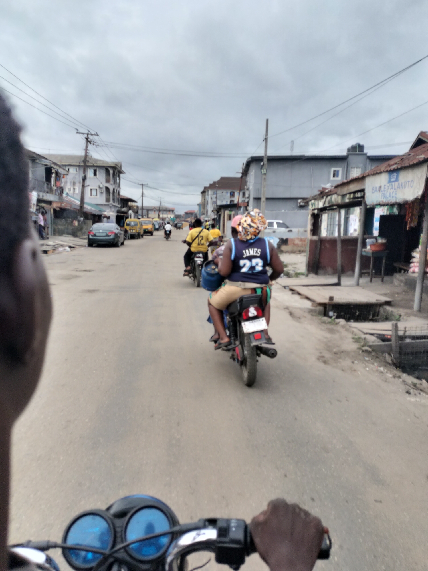 The image depicts a street scene in Alaiyabiagba, Nigeria, viewed from the perspective of a person riding a motorcycle. In front, another motorcycle carries two passengers, one wearing a blue "JAMES 23" jersey and a patterned headwrap. Further ahead, other motorcycles and vehicles, including yellow taxis, move along the street. Buildings line both sides of the road, with signs and structures suggesting commercial activity. The sky is overcast. The scene conveys a sense of urban activity and transport within the city.