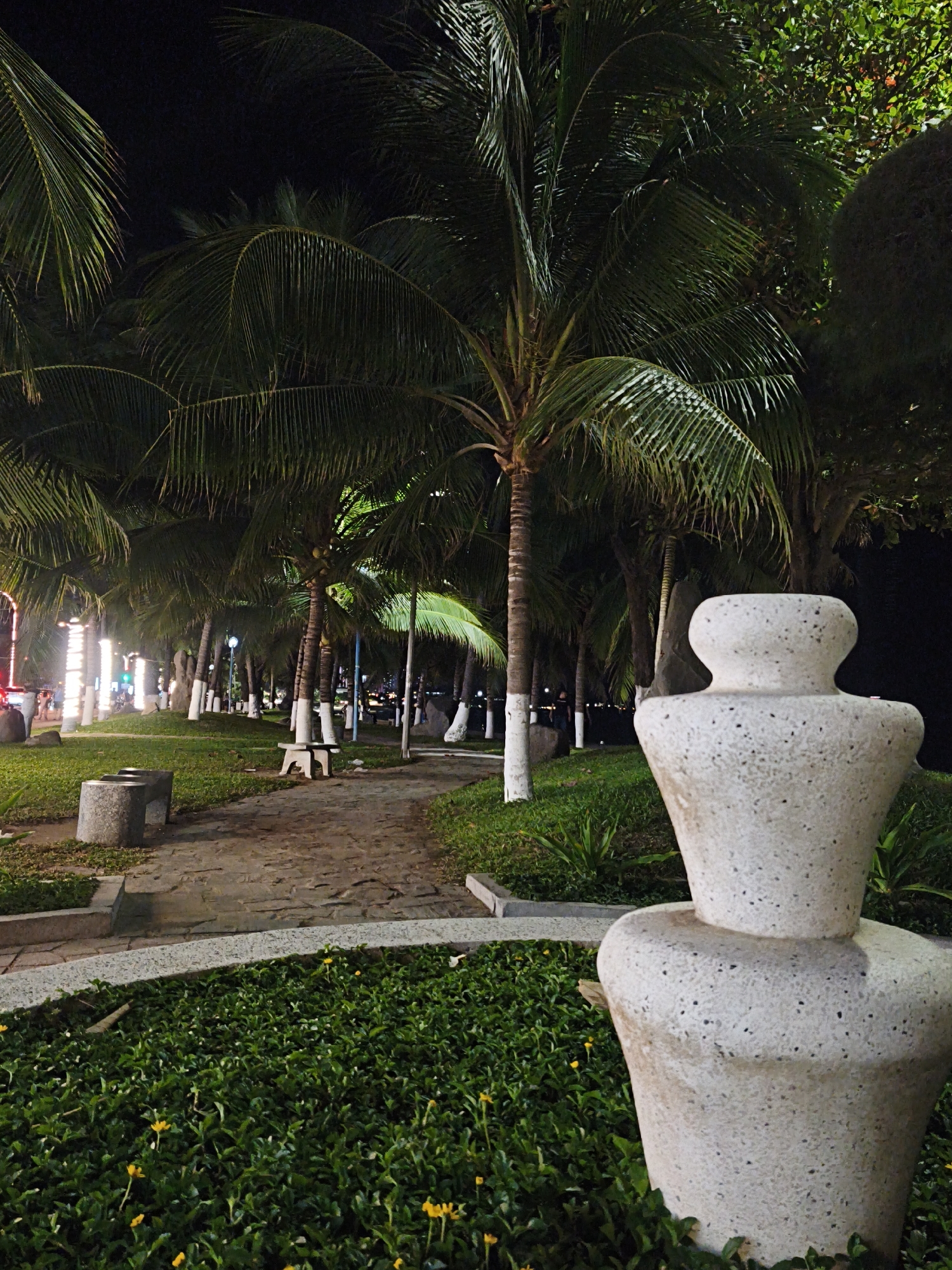 This image depicts a park scene at night in Nha Trang Ward, Vietnam. The foreground features a large, light-colored decorative stone vase with a textured surface, composed of two stacked sections. To the left of the vase, a cobblestone path leads into the background, bordered by a low concrete wall. Further left on the path are two stone benches. The path curves to the right, surrounded by lush green grass and small yellow flowers. A line of palm trees with white-painted trunks runs along the left side of the path, extending into the distance. In the background, beyond the palm trees, are more trees, some with visible foliage illuminated by artificial lights, suggesting a street or promenade with buildings and traffic. The overall atmosphere is one of a well-maintained urban park at night.