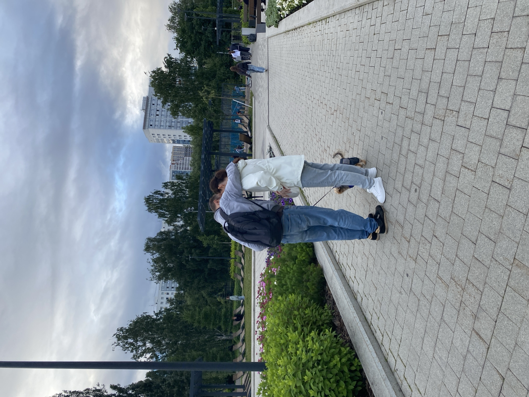 The image shows two people embracing in a park with a paved walkway. The woman is wearing a white jacket and gray pants, and the man is wearing a gray sweatshirt, blue jeans, and a black backpack. They are facing each other and appear to be in a romantic embrace. A small dog on a leash is visible to the right of the couple, wearing a black vest.
The park has trees and flower beds, with a building visible in the background. The sky is overcast, suggesting cloudy weather. The time of day appears to be late afternoon or early evening. The paved walkway is made of gray square tiles. In the background, other people can be seen walking in the park and on the walkway. There are also benches and playground equipment visible in the distance. The overall scene suggests a casual day spent in a city park in Perm, Russia.