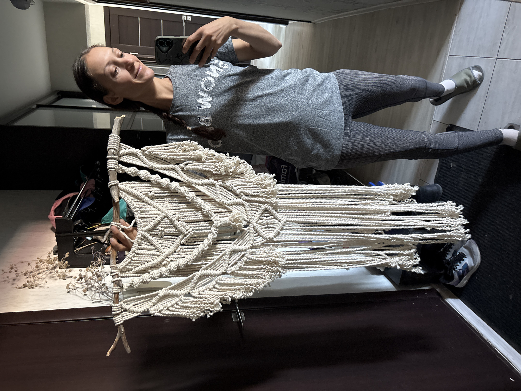 A woman with long brown hair in a braid holds up a macrame wall hanging. She wears a gray t-shirt and leggings, with light-colored socks and slip-on shoes. She is holding a cell phone, presumably taking a selfie. The background includes a hallway, tiled floors, and glimpses of interior spaces. The location appears to be an apartment. There is no definitive evidence to verify the city is Ulyanovsk, Russia.