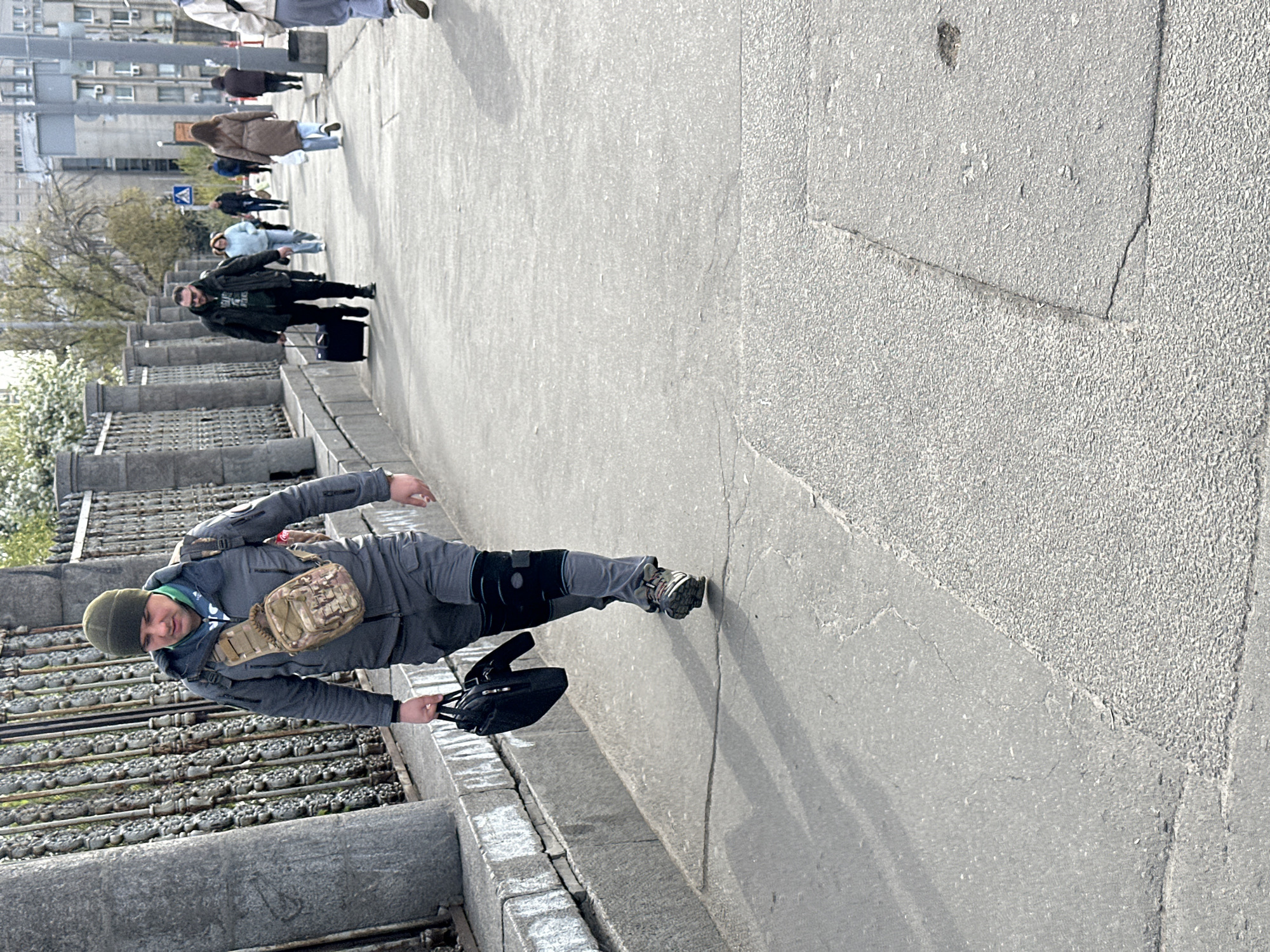 A man in a gray jacket, green hat, and camouflage bag is walking on a bridge in Kyiv, Ukraine. The bridge has decorative metal railings and concrete pillars. The man is also wearing a knee brace and carrying a black bag. Other pedestrians and buildings are visible in the background. The day is sunny.