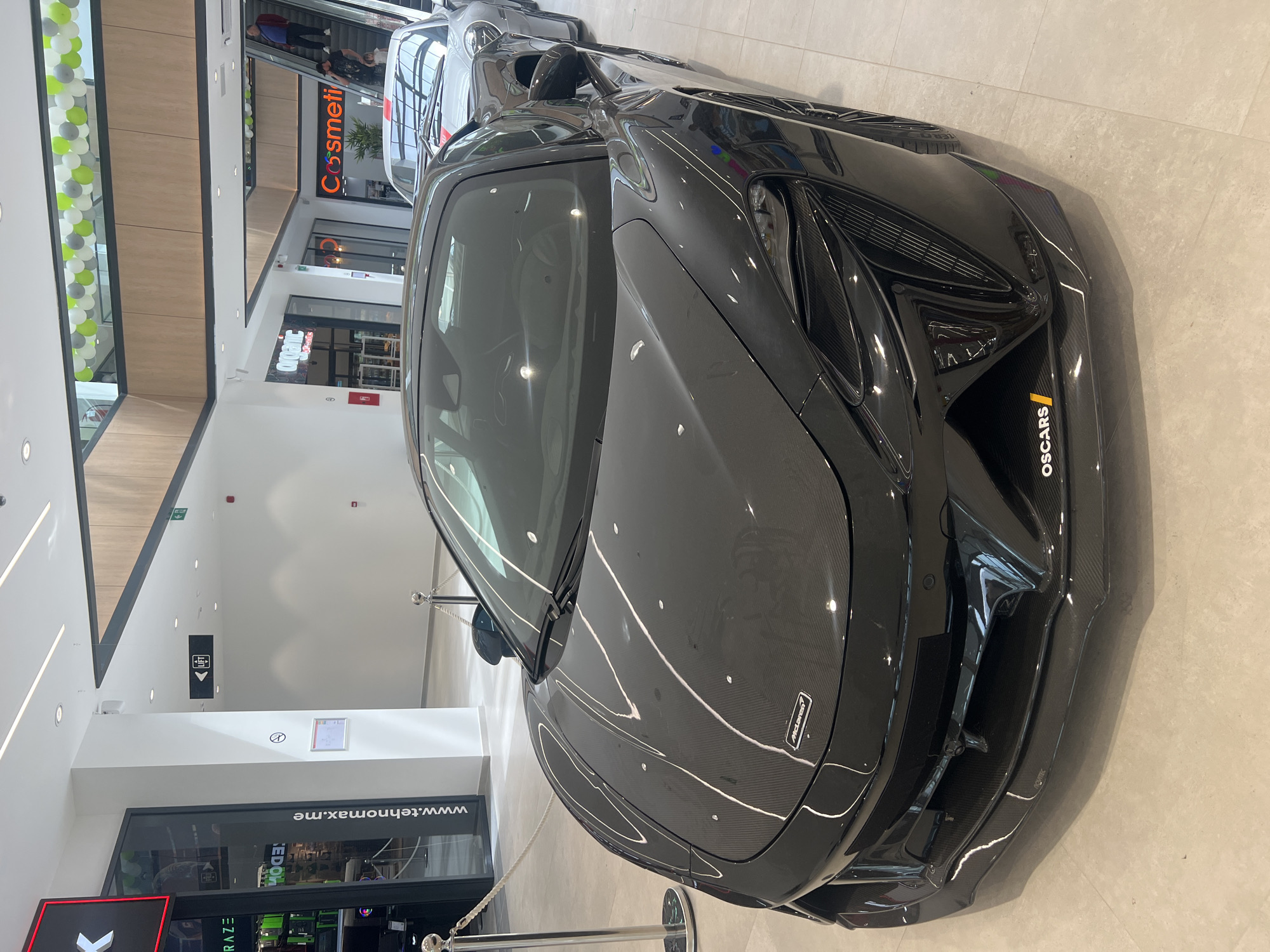 The image shows a black McLaren Artura car on display indoors, likely in a shopping mall or dealership in Bjeliši, Montenegro. The car is behind a silver stanchion and rope barrier. Other shops and stores are visible in the background. There are a few people visible in the background of the image. The scene is brightly lit with artificial light.