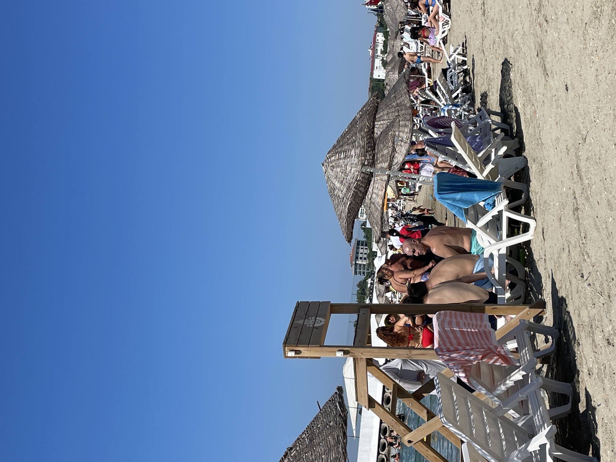 The image depicts a crowded beach scene under a clear blue sky, likely on a sunny day. In the foreground, numerous white plastic lounge chairs are arranged on the sand, some occupied by people and others draped with towels. Several thatched umbrellas provide shade over the lounge chairs. In the middle ground, a wooden structure with a sign featuring a hand symbol stands near the water's edge. People are visible wading and relaxing in the shallow water. Further back, more beachgoers are seen, as well as more umbrellas and buildings in the distance. The overall impression is a lively and popular beach resort, consistent with locations such as Caddebostan Beach in Kadıköy, Istanbul, Turkey, known for its sandy shores and recreational facilities.