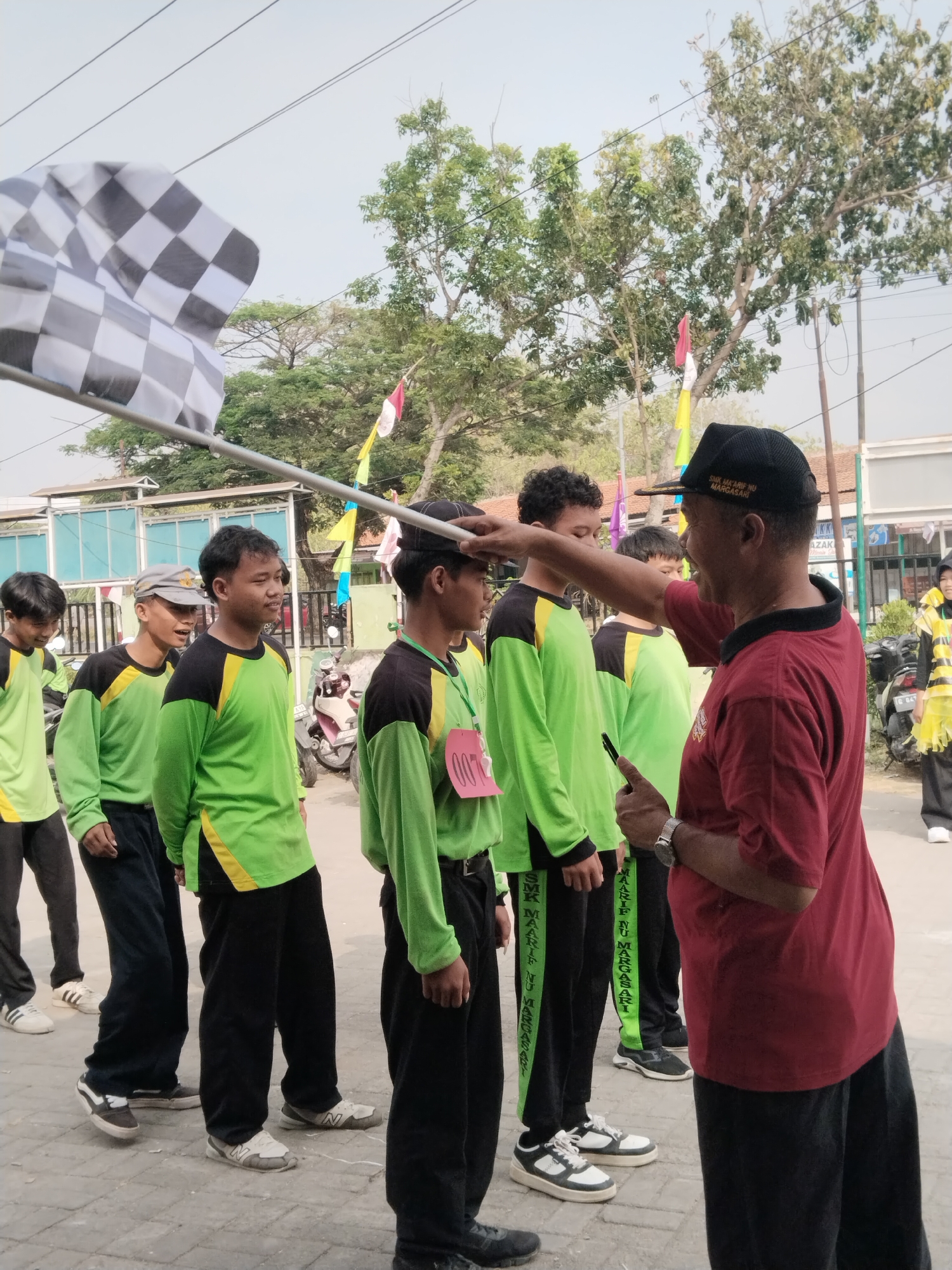 In the city of Pakulaut, Indonesia, a group of young males, dressed in matching green and black athletic shirts and black pants, stand in a line outdoors. A man wearing a red shirt and a black cap, with the words "SMK MA'ARIF NU MARGASARI" visible on it, is interacting with one of the young men, appearing to adjust something on his head. A checkered flag is held up in the foreground, suggesting the start or end of a race or competition. The background shows a street with trees, buildings, and parked motorcycles.