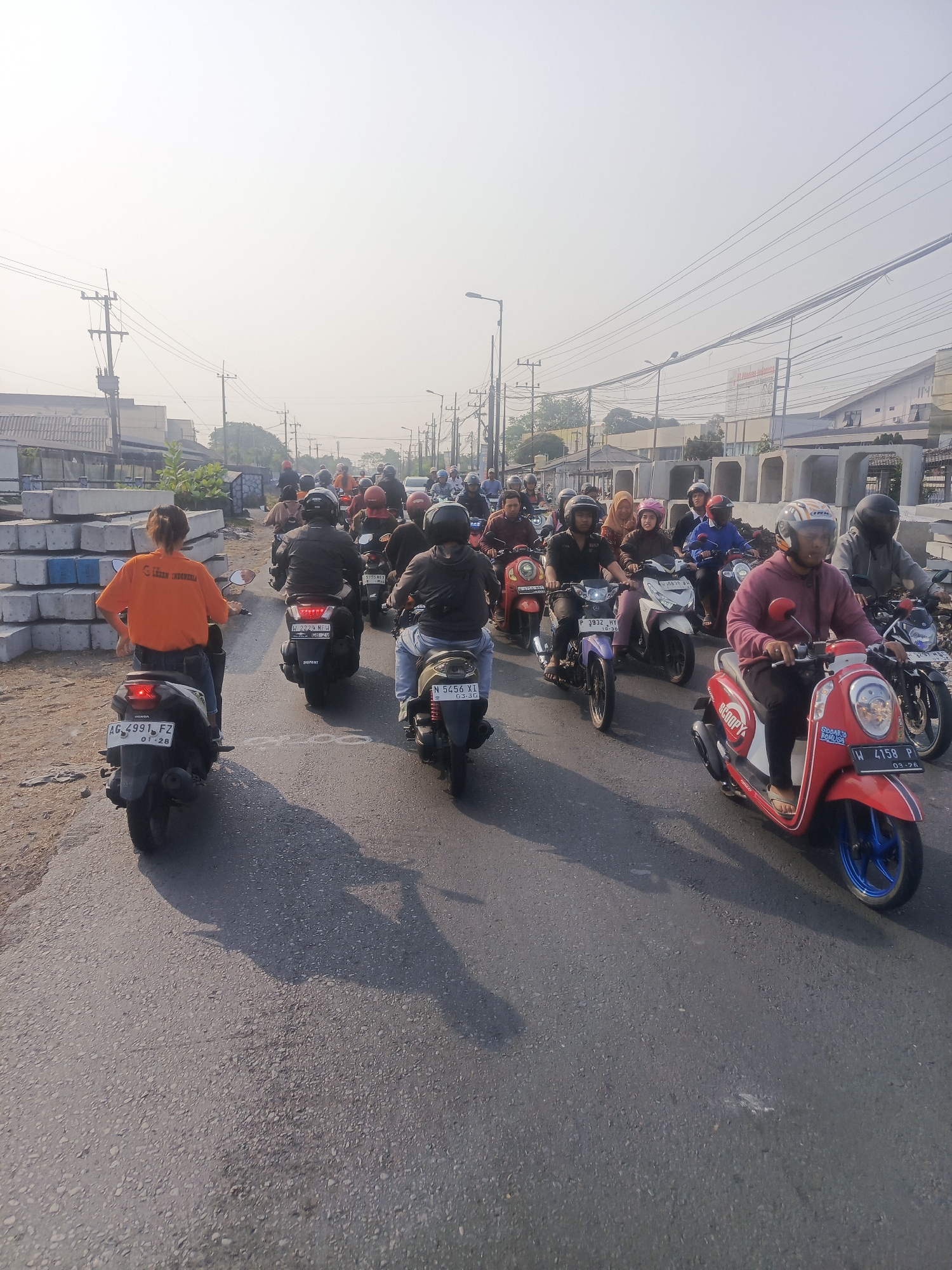 This is a forensic, matter-of-fact description of a street scene in Wedi, Indonesia.
The image depicts a road with a significant number of motorcycles, suggesting a traffic jam or congested area. The motorcycles are moving in the same direction, forming multiple lanes. Most of the riders are wearing helmets.
In the foreground, on the left side, a person wearing an orange shirt and blue jeans is riding a black scooter. The scooter has a license plate that reads "AG 4991 FZ" and a sticker with "01-28". The rider's back is to the camera.
Further back, there are numerous other motorcycles with riders. Some of the motorcycles are red, white, purple, and black. Several riders are visible wearing helmets of various colors, including red, blue, and black. Some passengers are also visible on the motorcycles.
The environment appears to be an urban or semi-urban area. On the left side of the road, there are large concrete blocks stacked, possibly indicating ongoing construction or materials storage. Utility poles with numerous wires are visible along the roadside, as are buildings in the background. The sky is bright and hazy, suggesting it is daytime, possibly early morning or late afternoon. The road surface is asphalt. The scene is consistent with a typical traffic situation in a bustling part of Wedi, Indonesia.