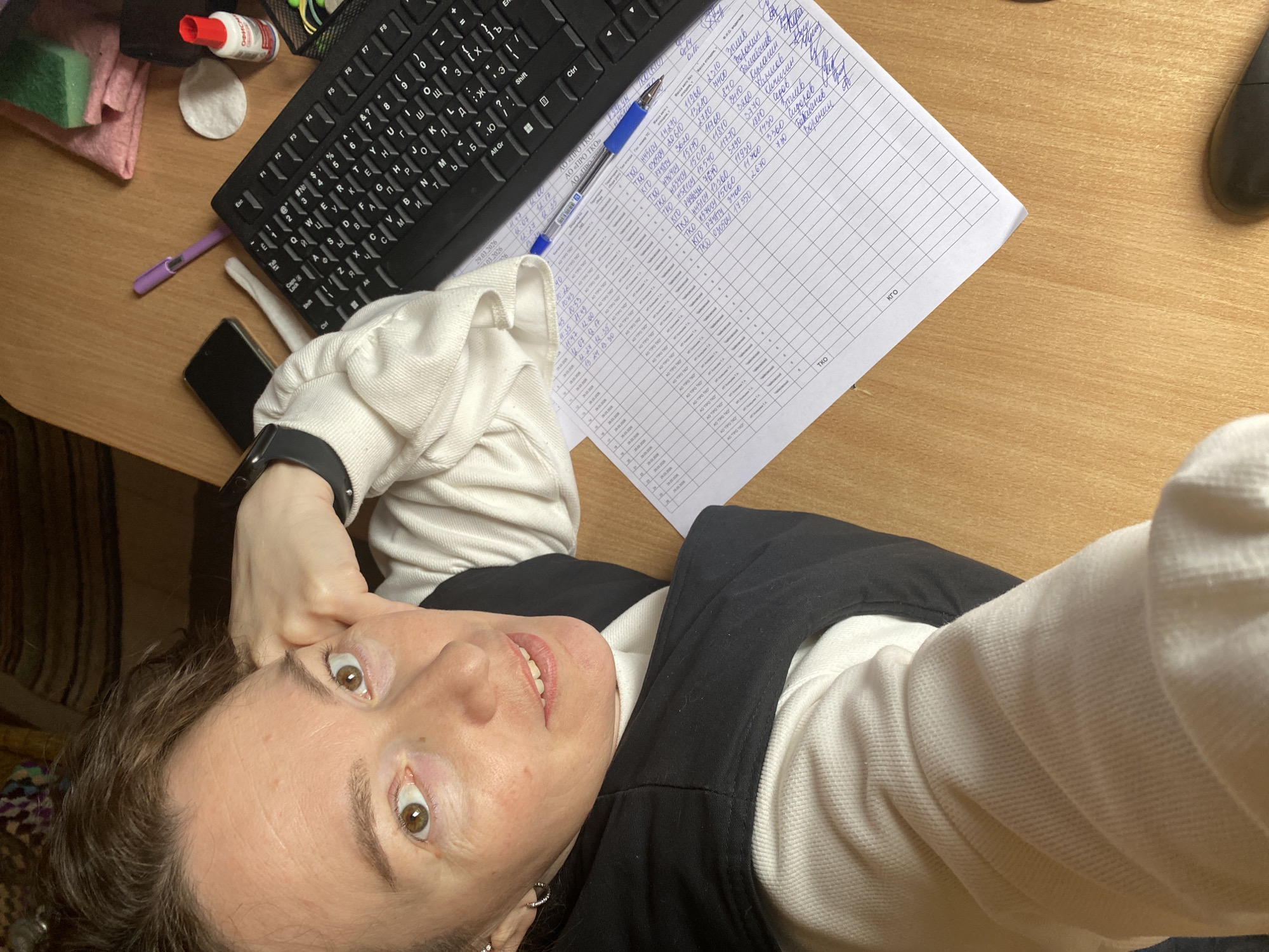 A person is taking a selfie with a desk in the background. On the desk are a black keyboard, a pen, a cell phone, cleaning supplies, and a sheet of paper filled with what appears to be handwritten Russian notes and gridlines. The person is wearing a white sweatshirt with a black vest over it. The location is potentially Ol'khovskoye, Russia, based on handwritten text on the sheet.
