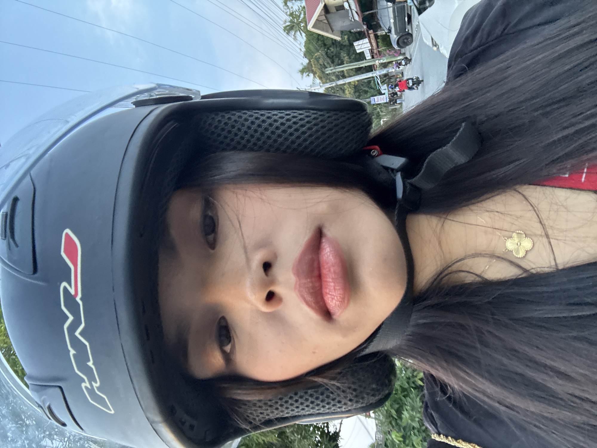 A medium shot depicts a woman wearing a black motorcycle helmet with a clear face shield. A motorcycle logo is visible on the left side of the helmet. The woman has long, straight, black hair and is wearing a black top with a gold necklace. She appears to be on a motorcycle, with a street scene visible in the background. In the background, there is a residential house with a red roof and white walls, power lines, palm trees, and other vehicles on the road. The picture was taken in Davao City, Philippines.