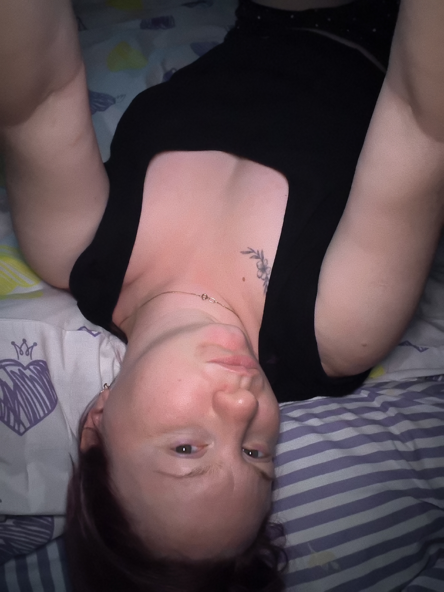 The image shows a person lying on a bed. The person is wearing a black top. The bed has striped sheets and pillowcases. There is a tattoo of a flower on the person's chest. The person's face is partially visible. The background is blurred. The image is taken from a top-down perspective. The location is not specified in the image.