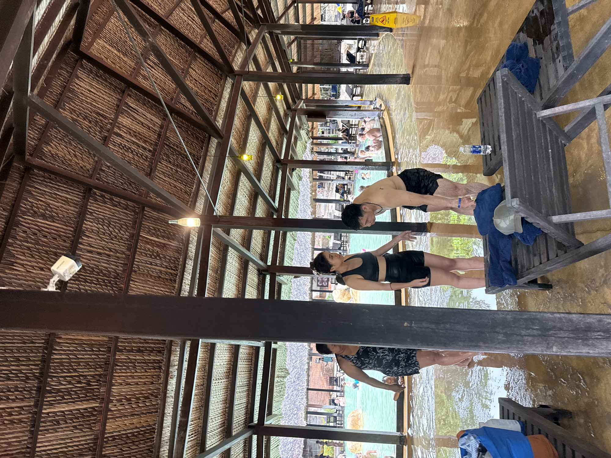The image shows an indoor pool area with a thatched roof supported by wooden beams and columns. There are several people in swim attire in and around the pool. In the foreground, two women and one man are standing in shallow water. The woman on the left is wearing a black and white patterned swimsuit, the woman in the center is wearing a black sports bra and black shorts, and the man on the right is wearing black swim trunks. A dark blue tote bag with clothes and a water bottle are on a wooden lounge chair to the left. Another lounge chair with blue towels and a hat is in the center. In the background, more people are visible in the pool and relaxing on lounge chairs. The floor is wet and appears to be made of smooth, light-colored material. The overall environment suggests a resort or spa with a tropical theme. The location is identified as a popular hot spring resort in Nha Trang, Vietnam.