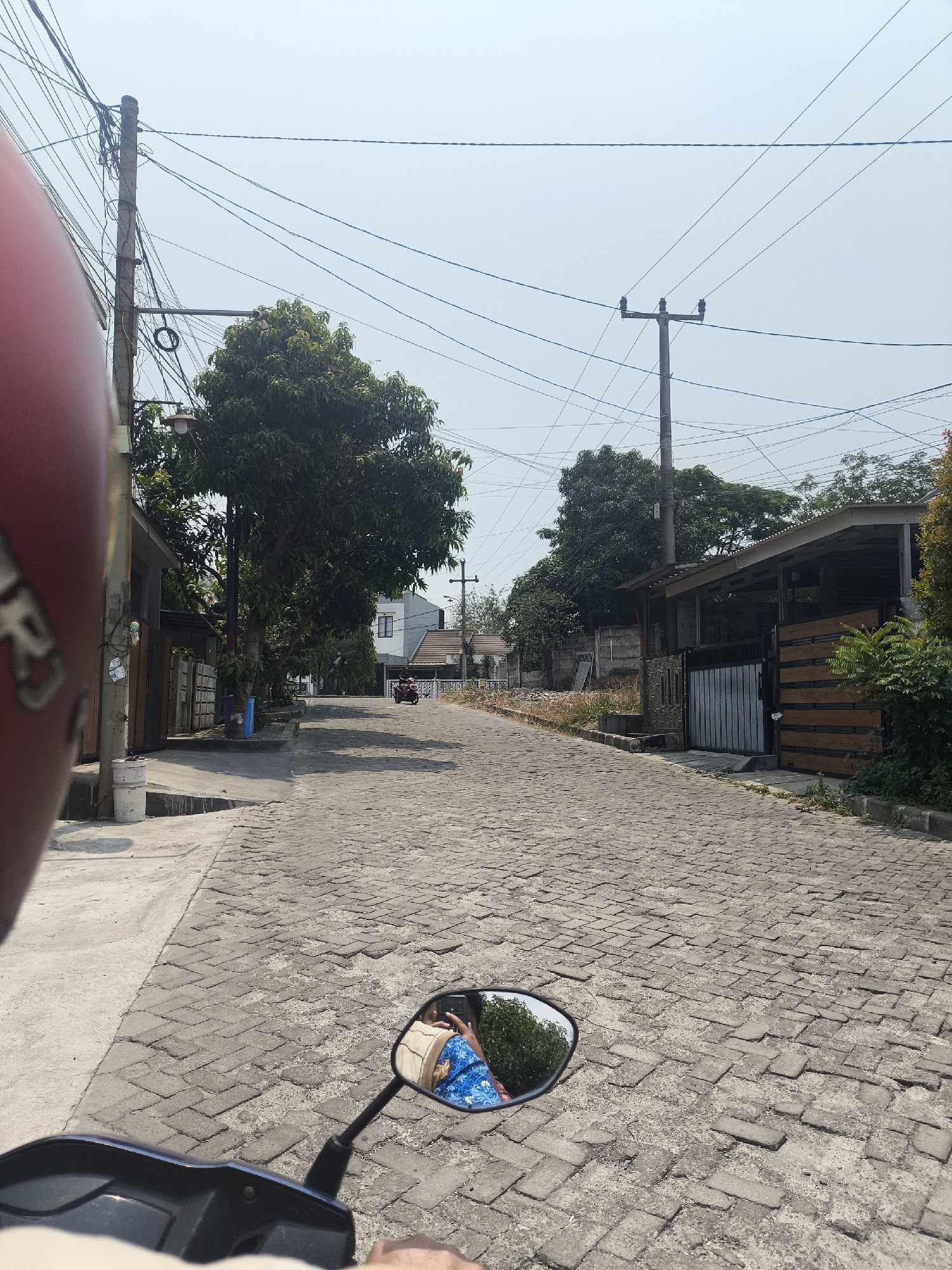 The image captures a perspective from a motorcycle traveling on a paved road in Cilegon, Indonesia. The road is constructed from gray interlocking bricks, with visible wear and some unevenness. The scene is bordered by houses, some with simple fences and gates, and lush green trees. Utility poles with numerous electrical wires stretch across the sky, which is a hazy light blue. In the foreground, the red helmet of the motorcycle rider is partially visible on the left. A side mirror reflects the rider wearing a blue and white patterned shirt and holding a mobile phone. Further down the road, another motorcycle is seen in motion. The environment suggests a residential area in a warm, possibly humid climate.